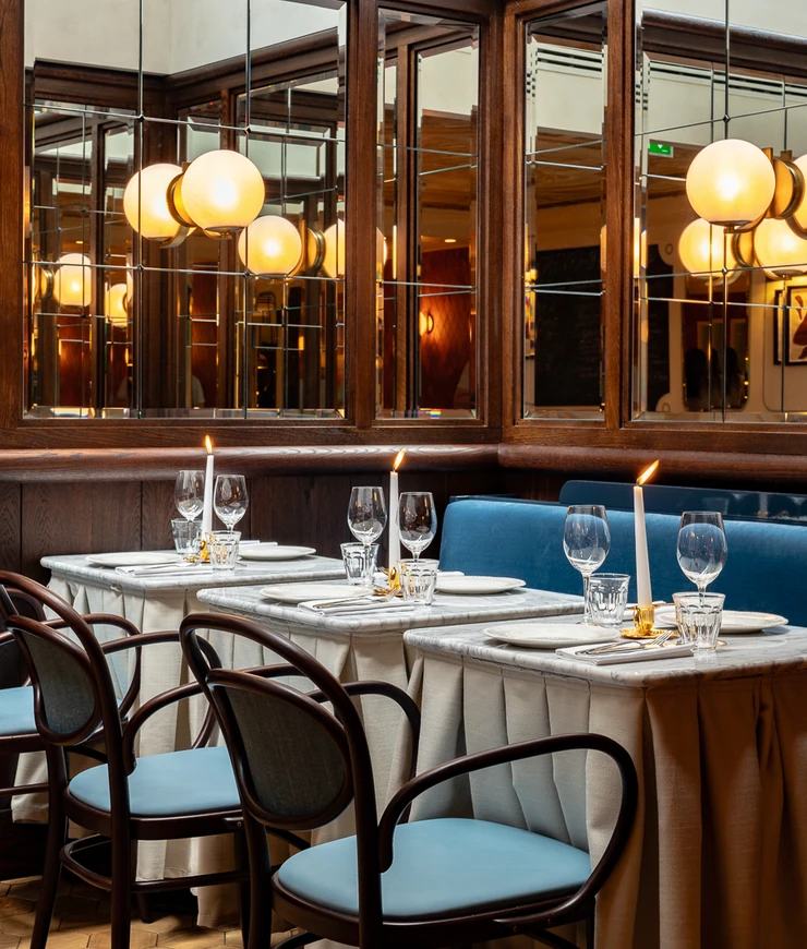 Des tables de restaurant élégantes avec des nappes blanches, des chaises rembourrées bleues, des bougies allumées, de la verrerie et des miroirs reflétant un éclairage chaleureux.