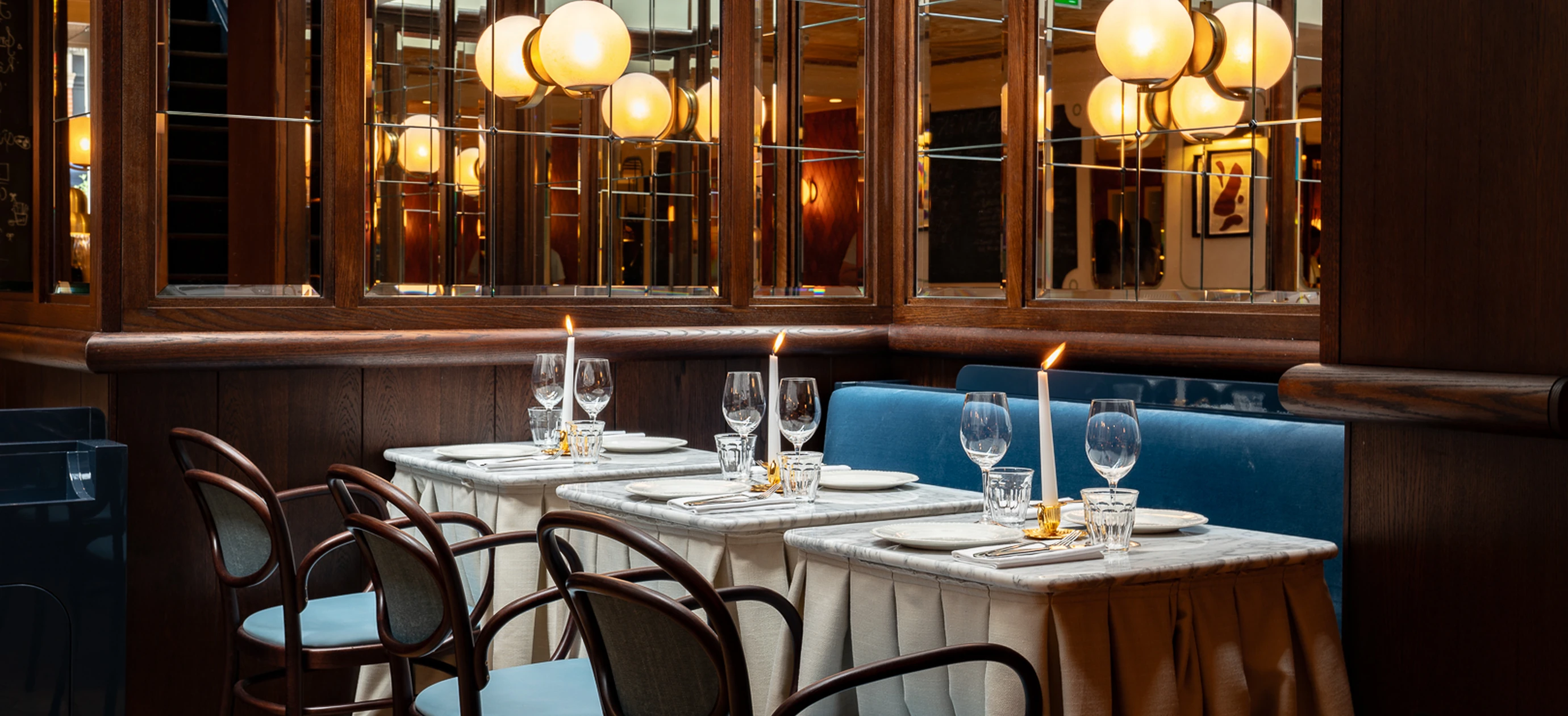 D'élégantes tables de restaurant avec des bougies blanches, de la verrerie et des assiettes sont disposées à côté d'un banc rembourré bleu, sous des appliques murales rondes et des miroirs.