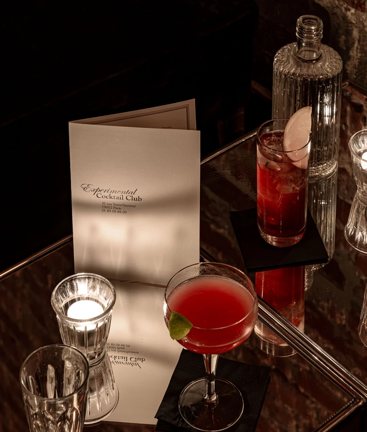 Two cocktails on a glass table at Experimental Cocktail Club; one red cocktail in a coupe glass with a lime wedge, the other in a highball glass with a lemon slice, accompanied by a small lit candle and a folded menu.
