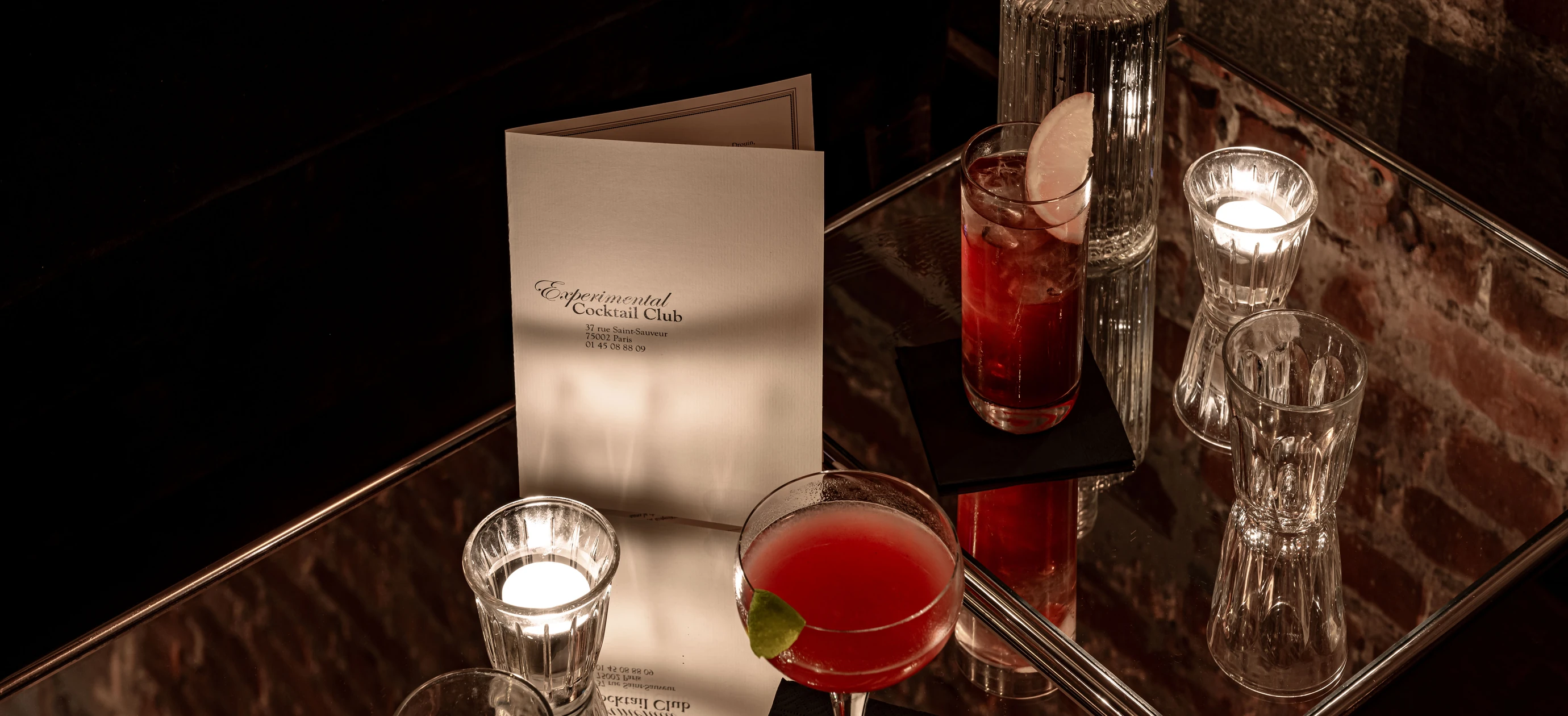 Two cocktails on a reflective glass table with candles and a menu from Experimental Cocktail Club in a dimly lit brick-walled setting.
