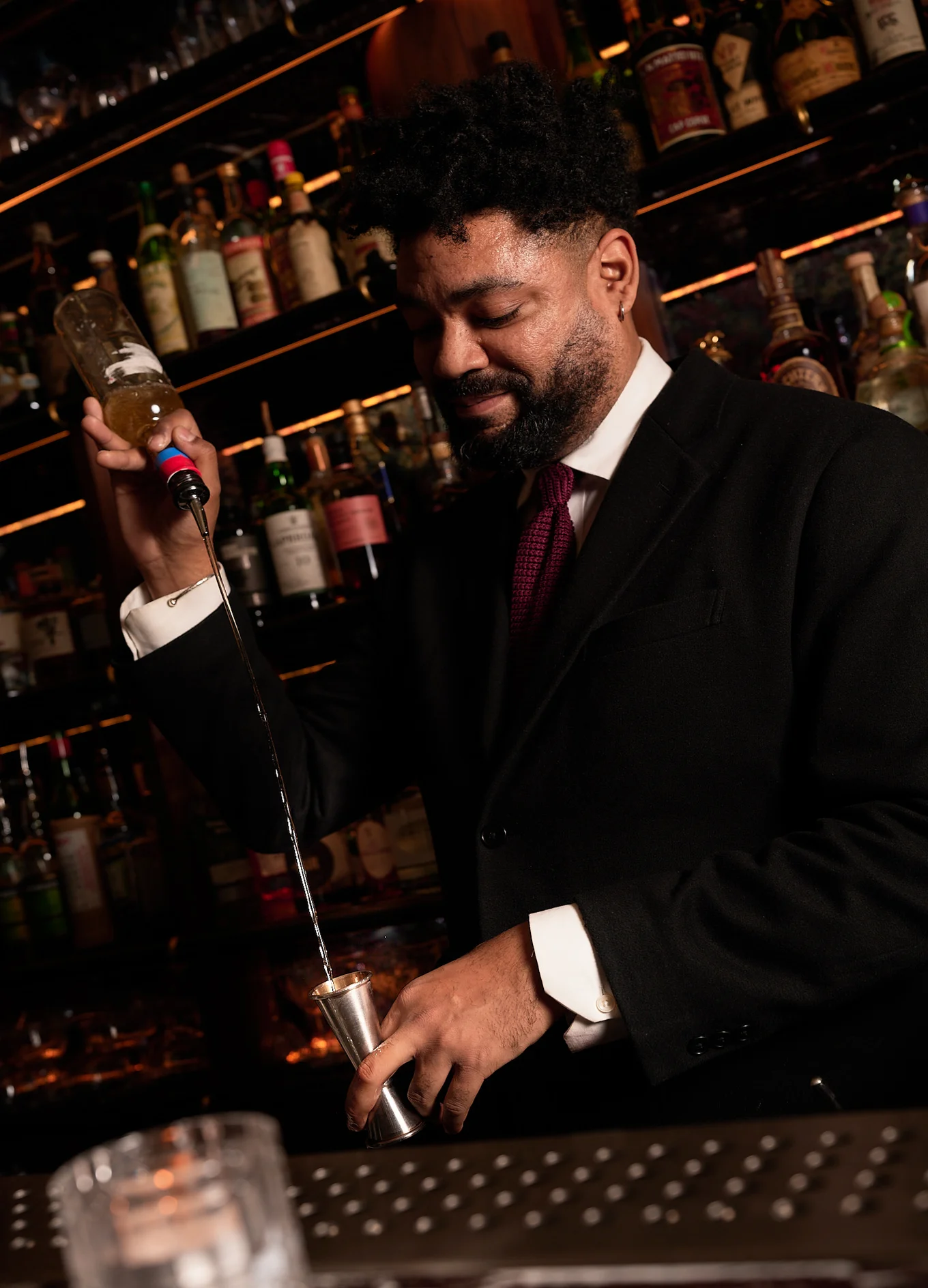 Bartender in a black suit and red tie pouring liquor from a bottle into a jigger in a dimly lit bar.