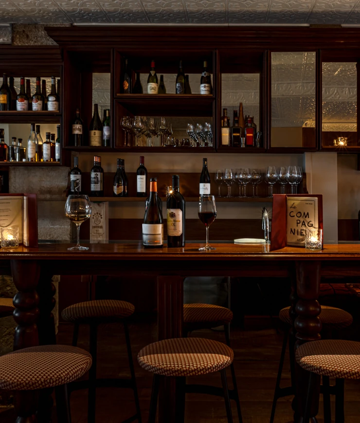 Cozy bar with wooden shelves holding wine bottles and glasses, a wooden counter with bottles, wine glasses, a lit candle, and cushioned stools.