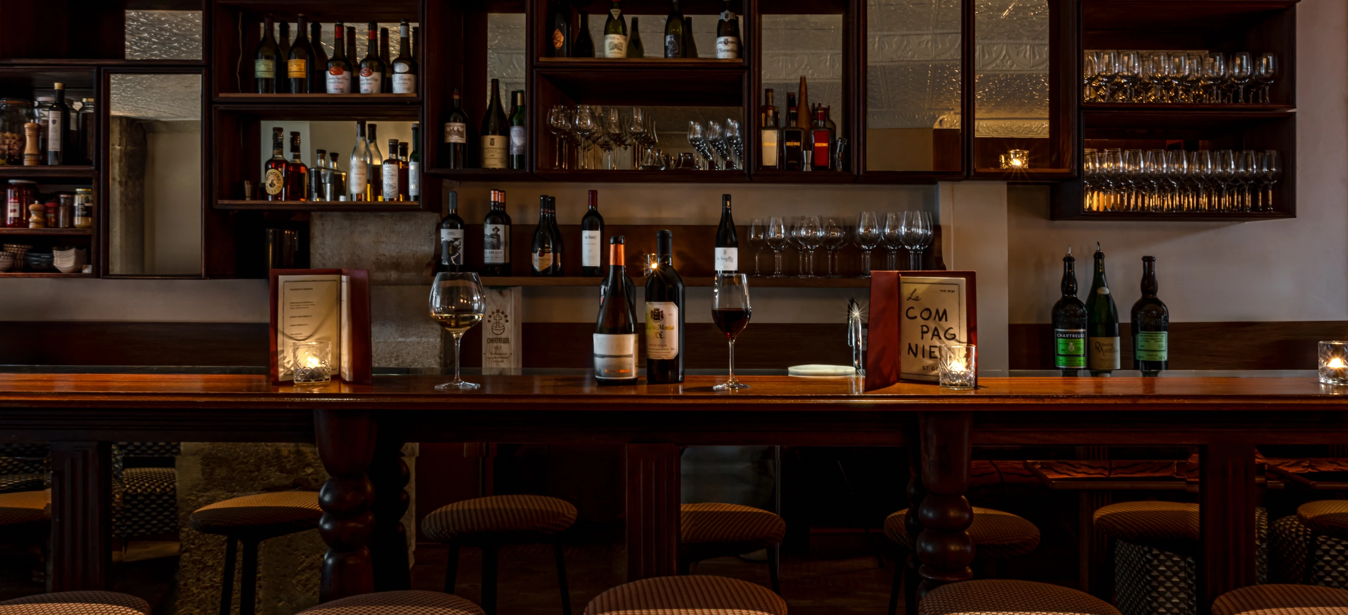 Cozy bar interior with wooden counter, wine bottles, wine glasses, menus, and soft candle lighting.