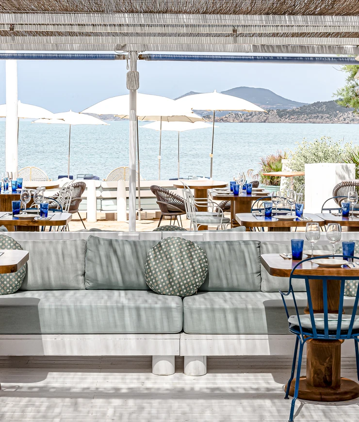 Área de comedor al aire libre junto a la playa con asientos acolchados, mesas de madera, vasos azules, sombrillas blancas y vista al mar con colinas distantes.