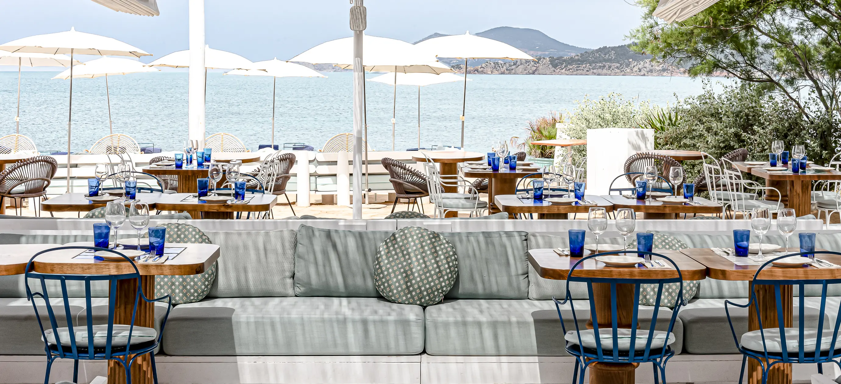 Restaurante al aire libre junto a la playa con mesas de madera, sillas azules, sombrillas y vistas al mar y a las colinas lejanas.