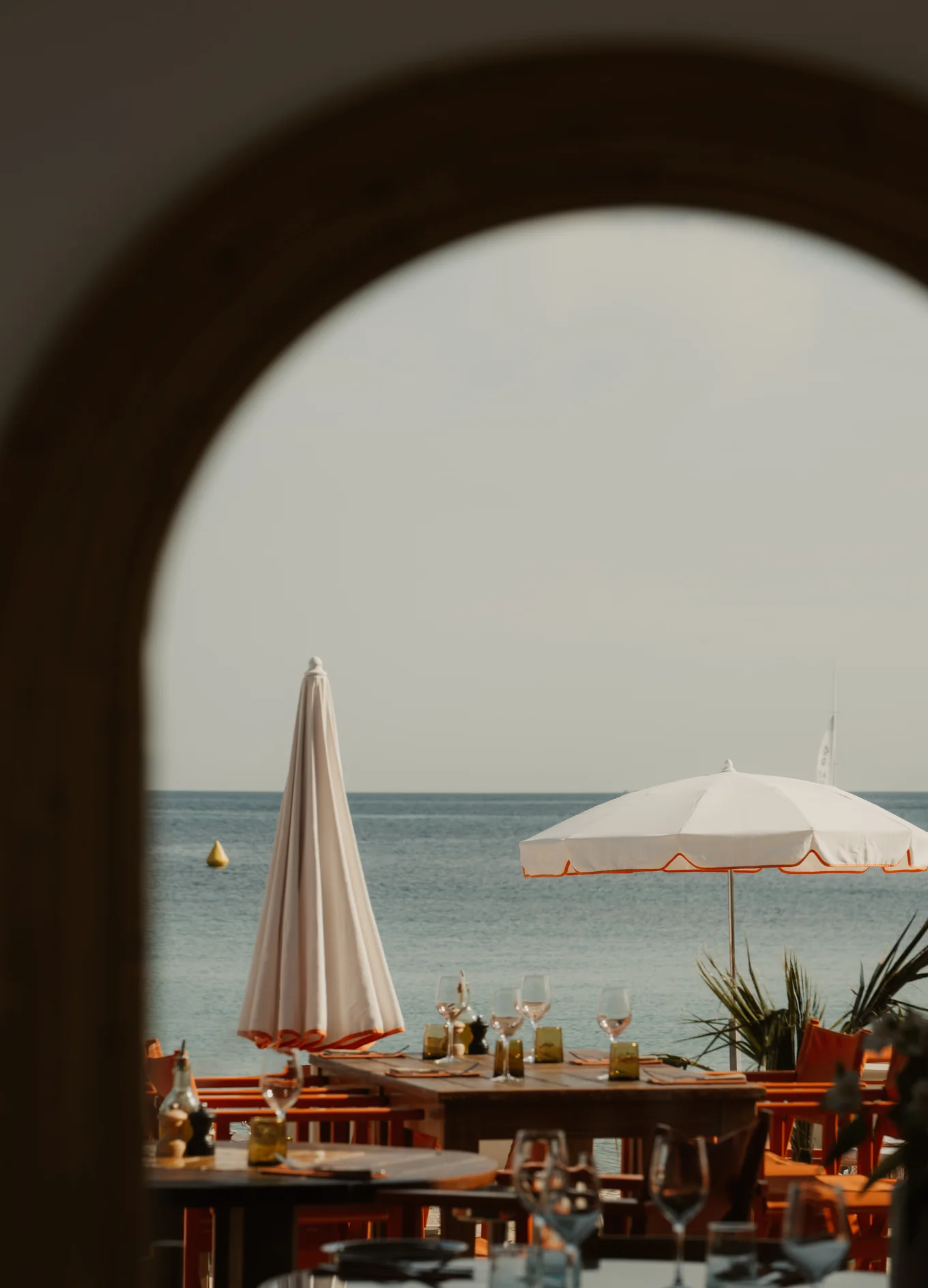 Outdoor beachside dining area with wooden tables, orange chairs, white umbrellas, and view of calm sea.