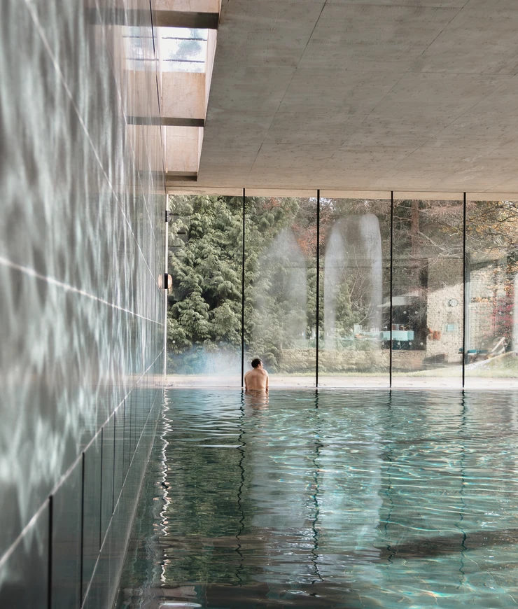 Person sitting alone in an indoor pool facing large glass windows with a view of trees and outdoor scenery.