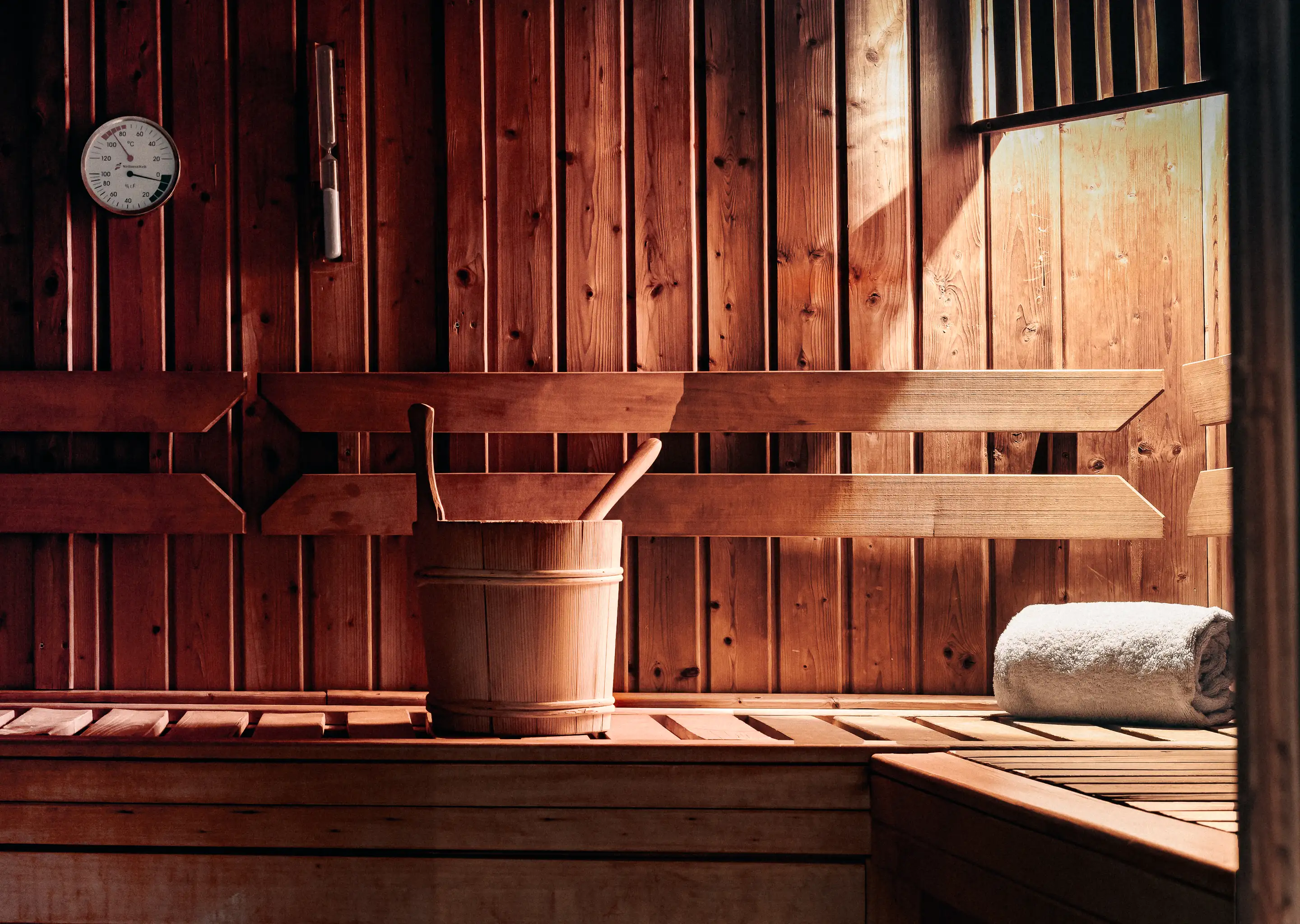 Wooden sauna interior with bench, bucket and ladle, white towel, and a thermometer on the wall.