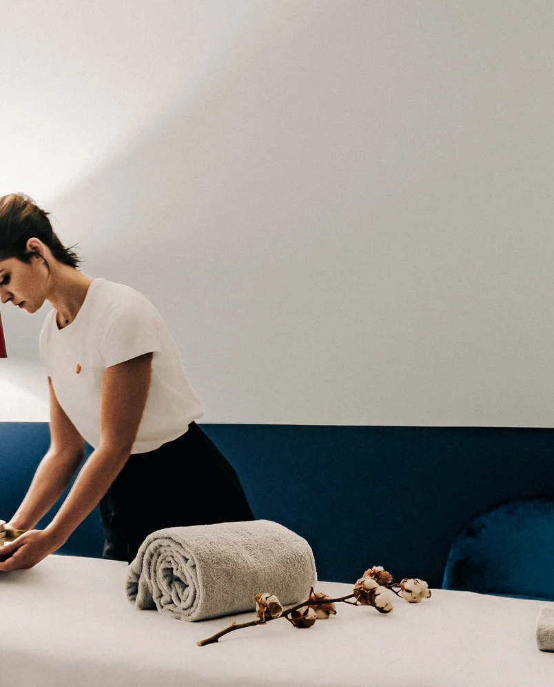 A woman in a white shirt preparing a massage table with a rolled towel and a cotton branch in a spa room.