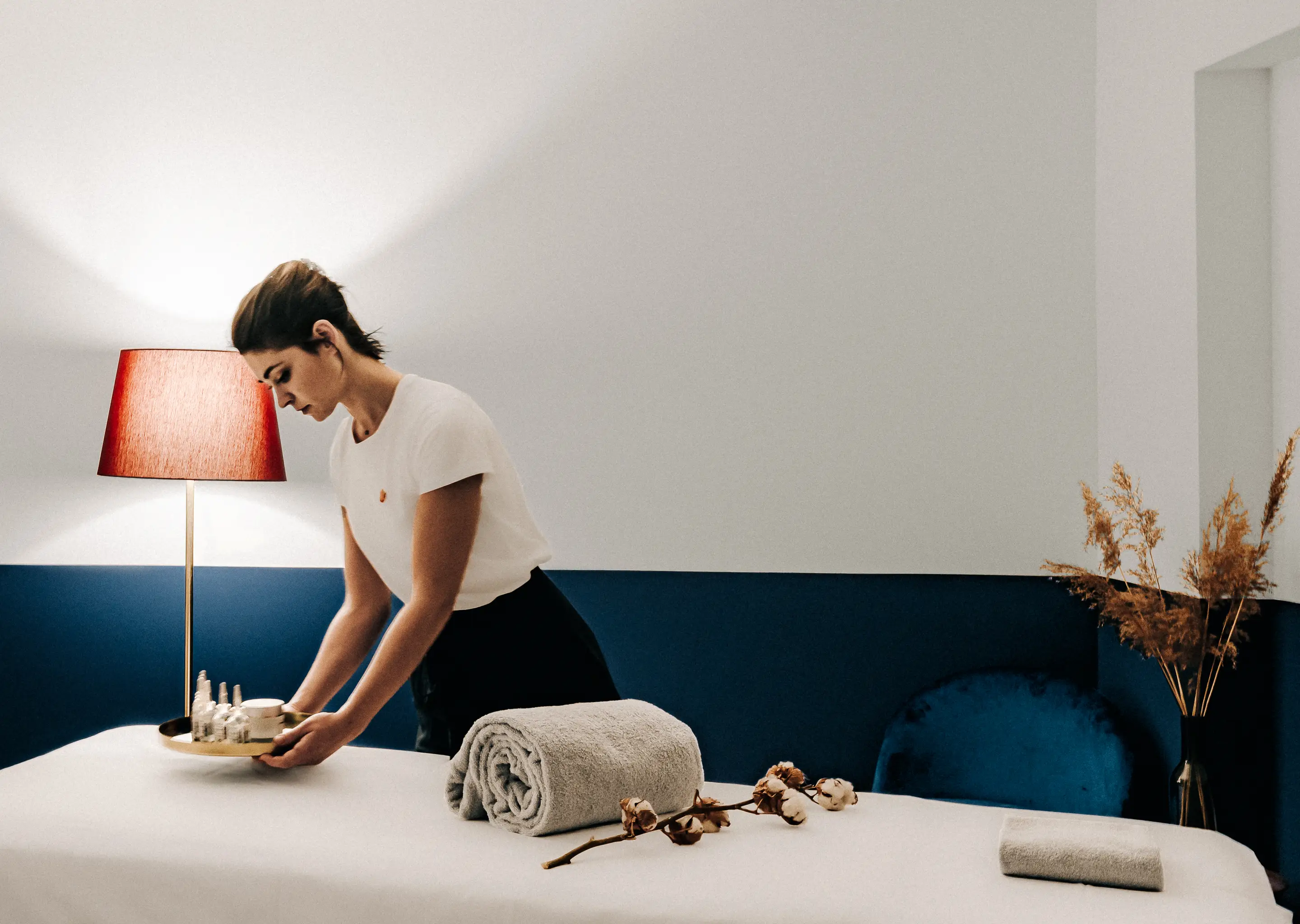 Woman arranging spa products on a massage table in a modern room with a red lamp and dried plants.
