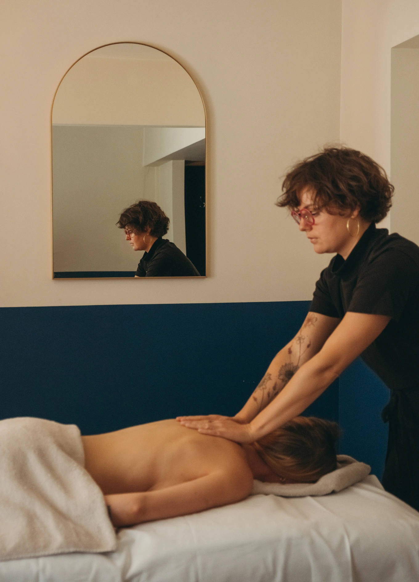 Therapist with short curly hair and glasses giving a back massage to a person lying face down on a massage table covered with a towel.