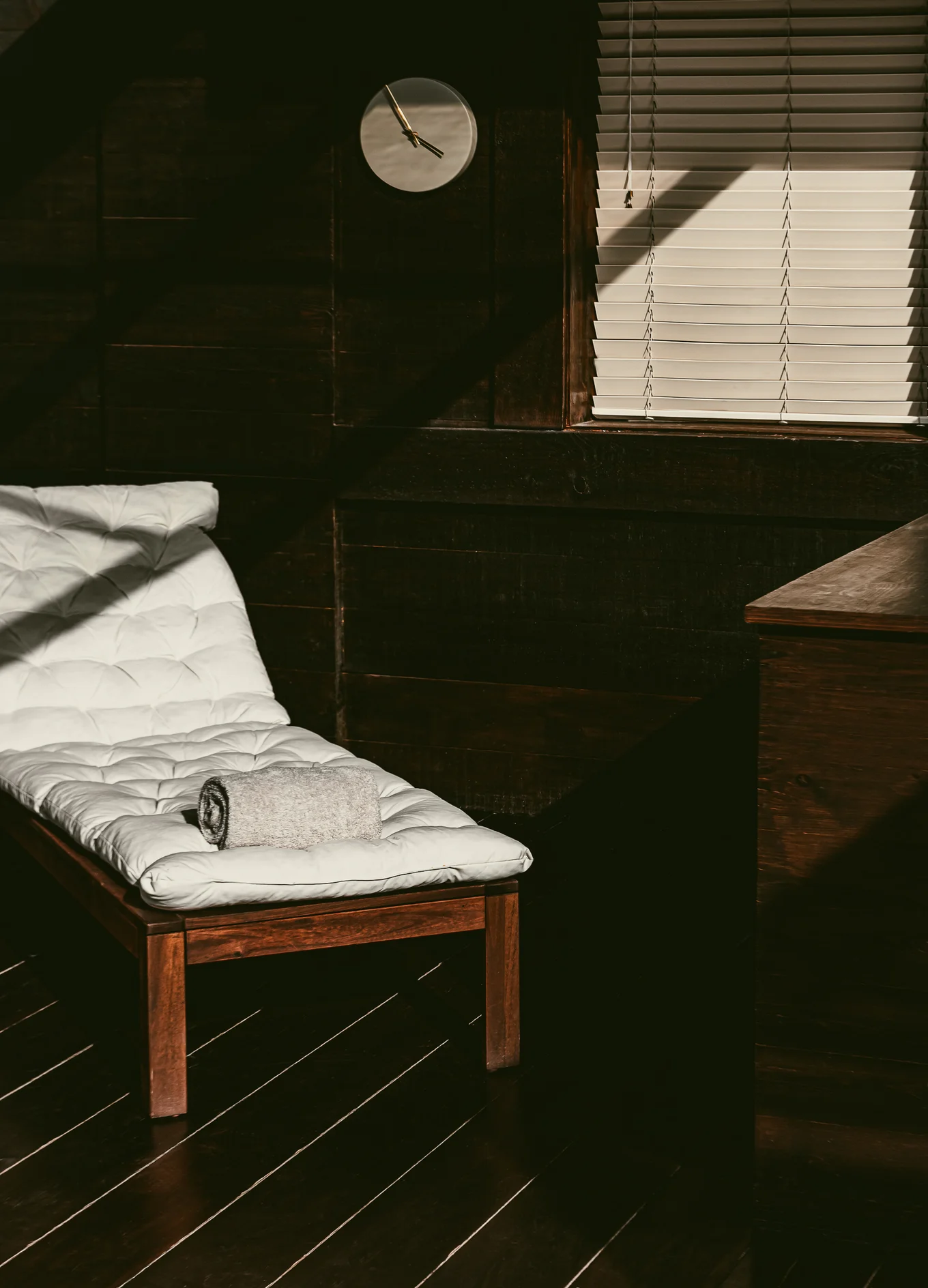 Wooden lounge chair with a white cushion and a rolled gray towel placed on it, next to a wooden wall with blinds partially closed and a round white clock showing 10:10.