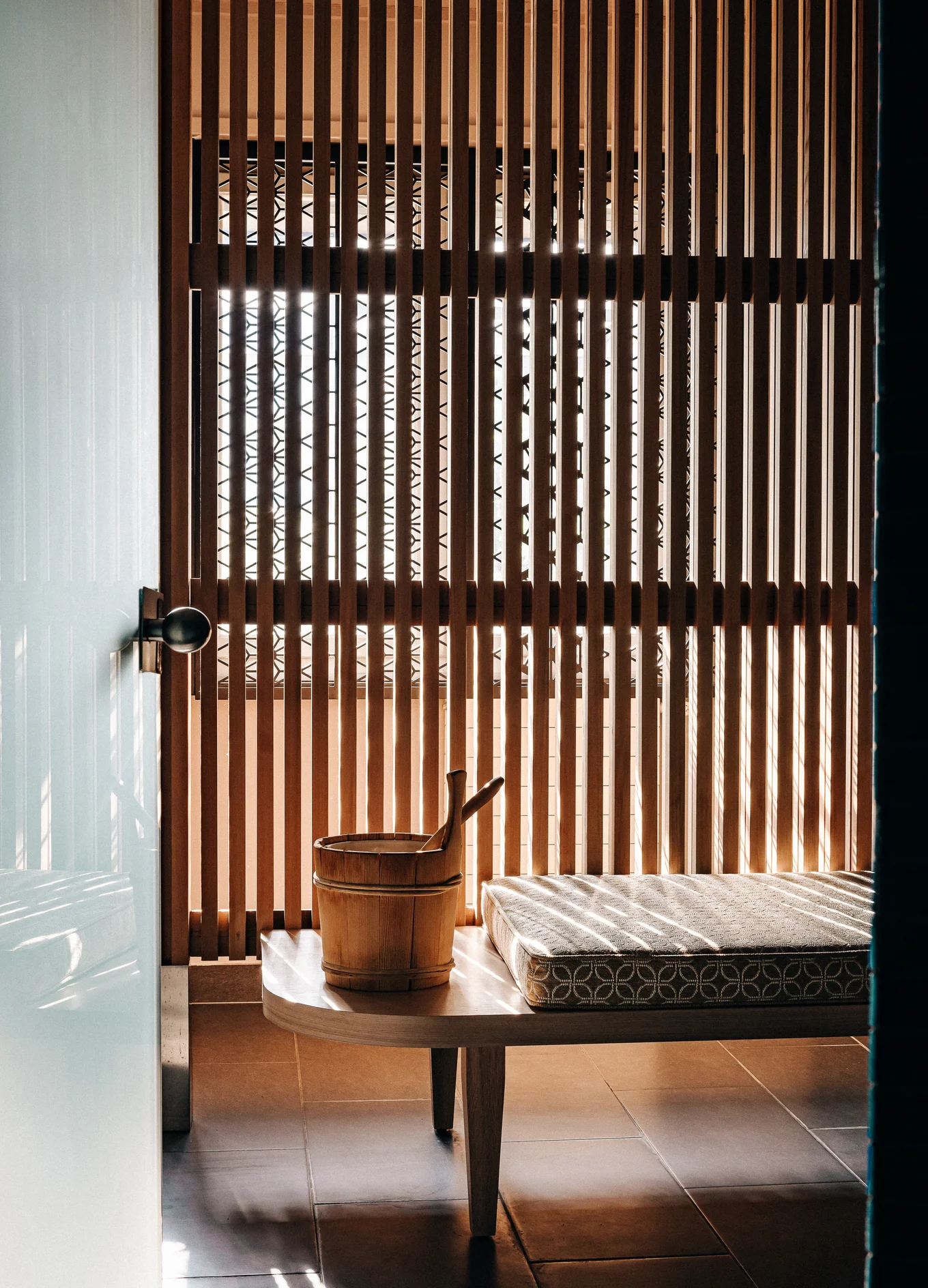 Wooden bench with cushion and a wooden bucket with ladle in a sunlit room with vertical wooden slat walls.