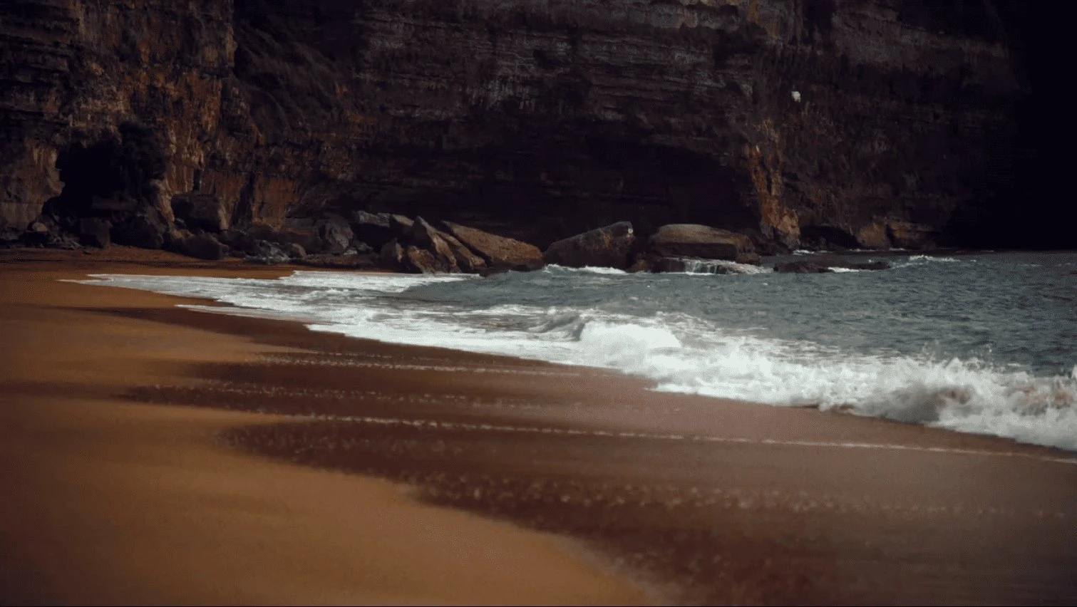 Waves gently crashing onto a sandy beach with rocky cliffs in the background.