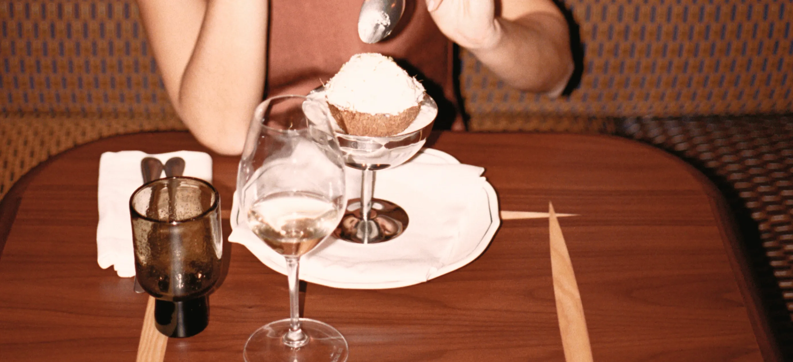 Person serving a dessert in a glass bowl topped with shredded coconut on a wooden table with wine glasses and cutlery.