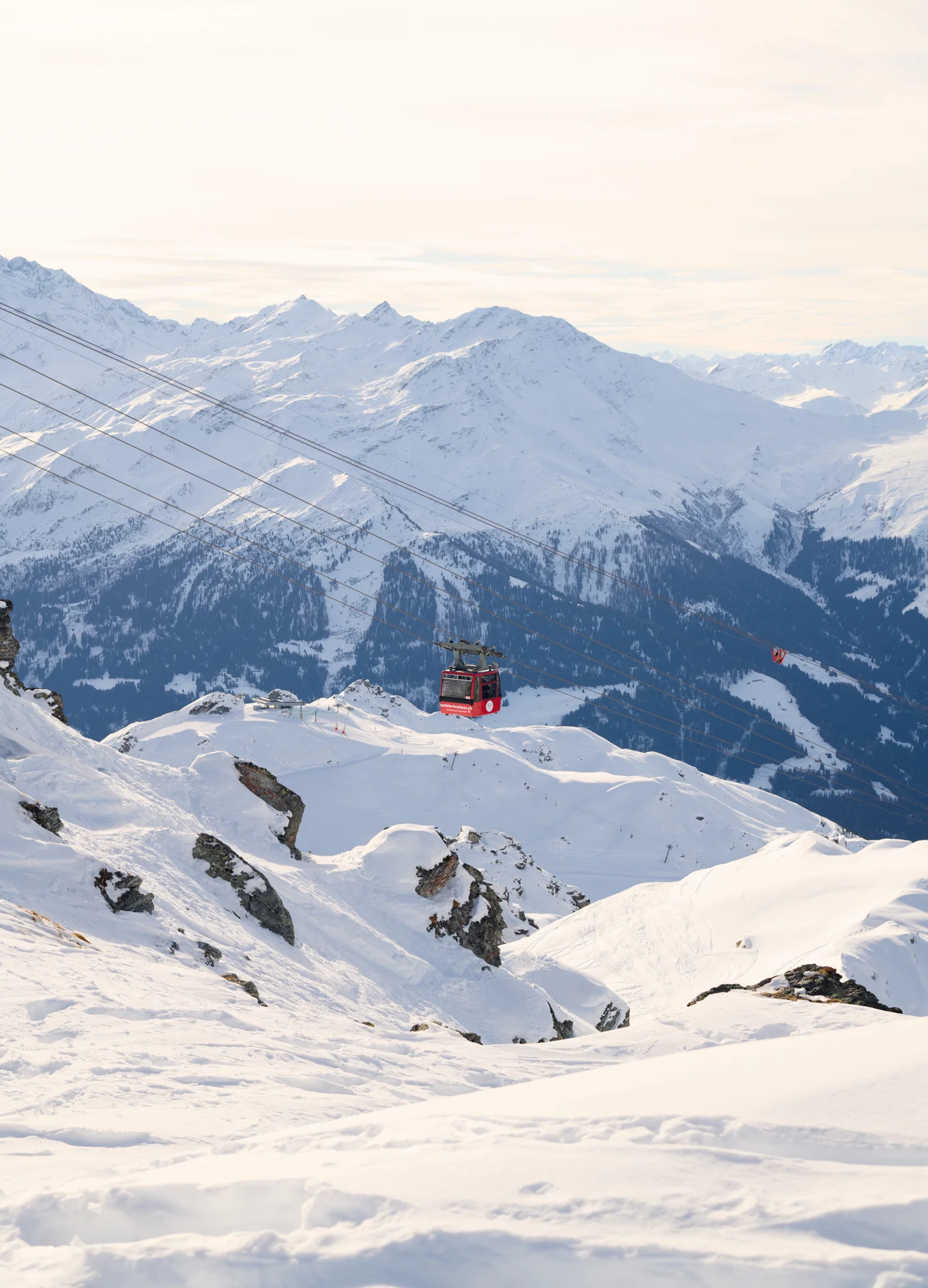 Red cable car suspended on wires over snow-covered mountains with distant snowy peaks under a cloudy sky.