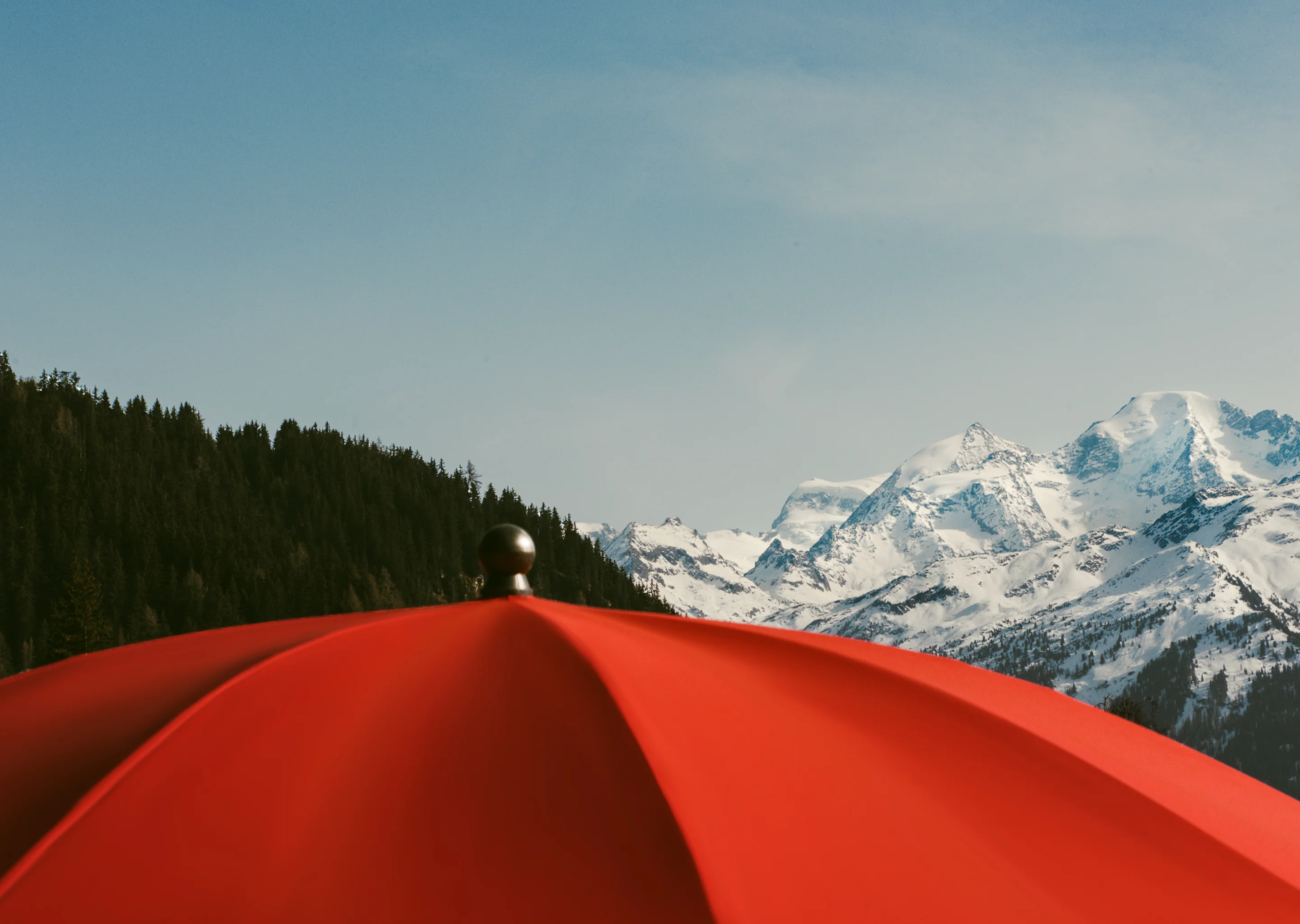 Top of a red umbrella with snow-covered mountains and pine trees in the background under a clear sky.