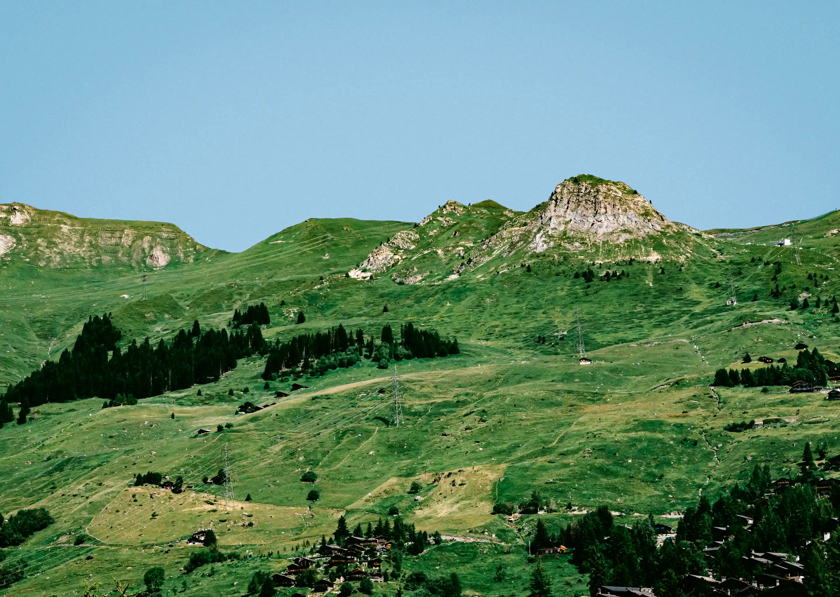 Green mountainous landscape with scattered small houses and trees under a clear blue sky.