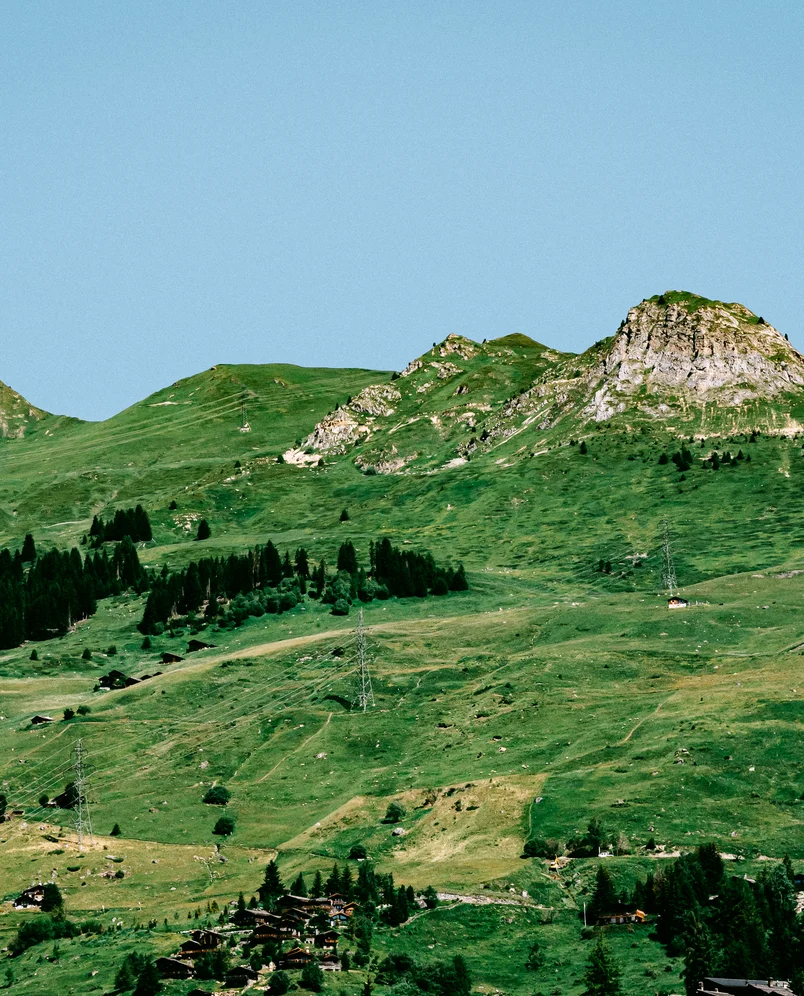 Green mountainous landscape with scattered trees, small buildings, and a clear blue sky.