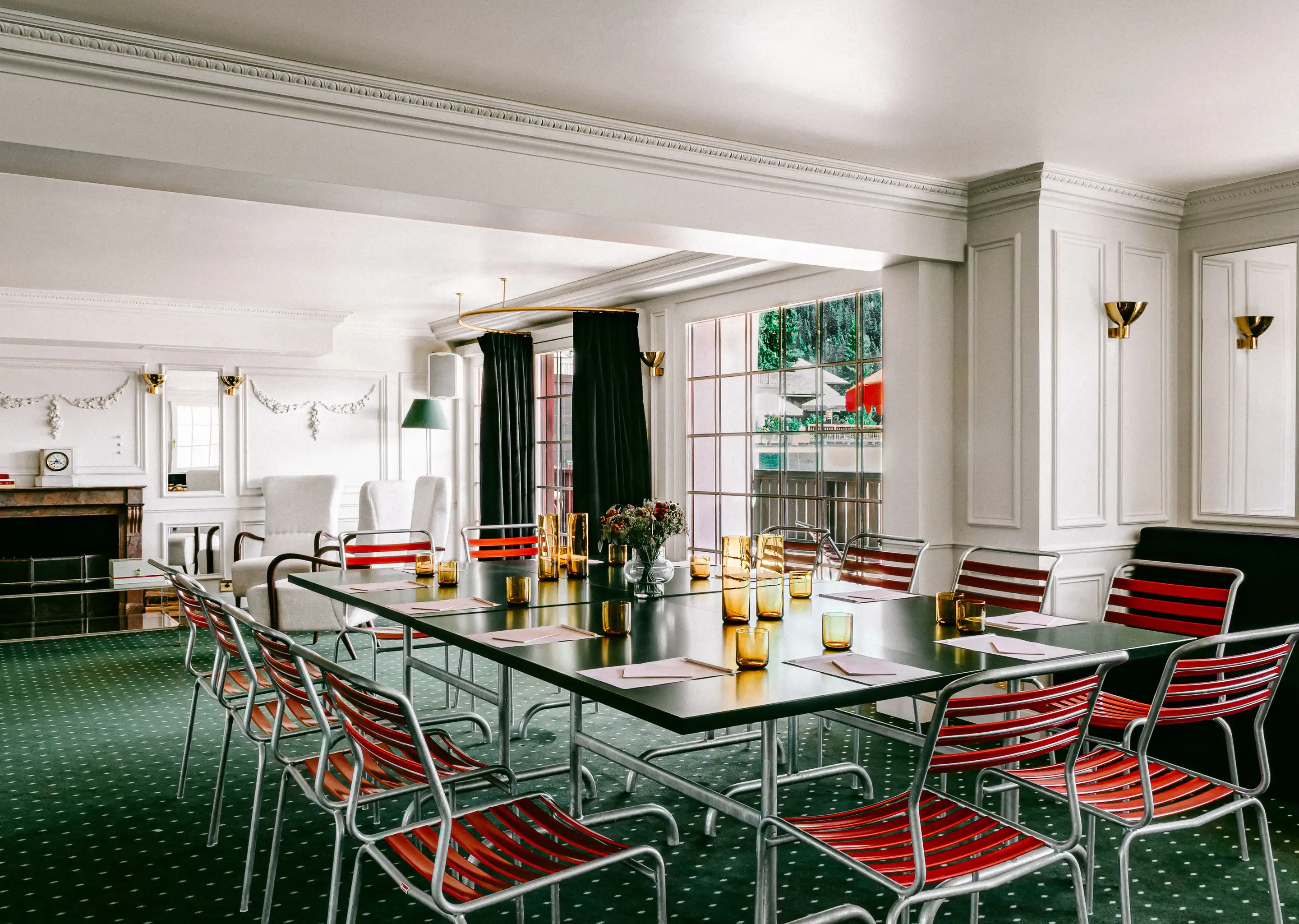 Elegant conference room with a rectangular black table surrounded by red and silver chairs, amber glassware, and white notepads, set in a room with white paneled walls and green carpet.