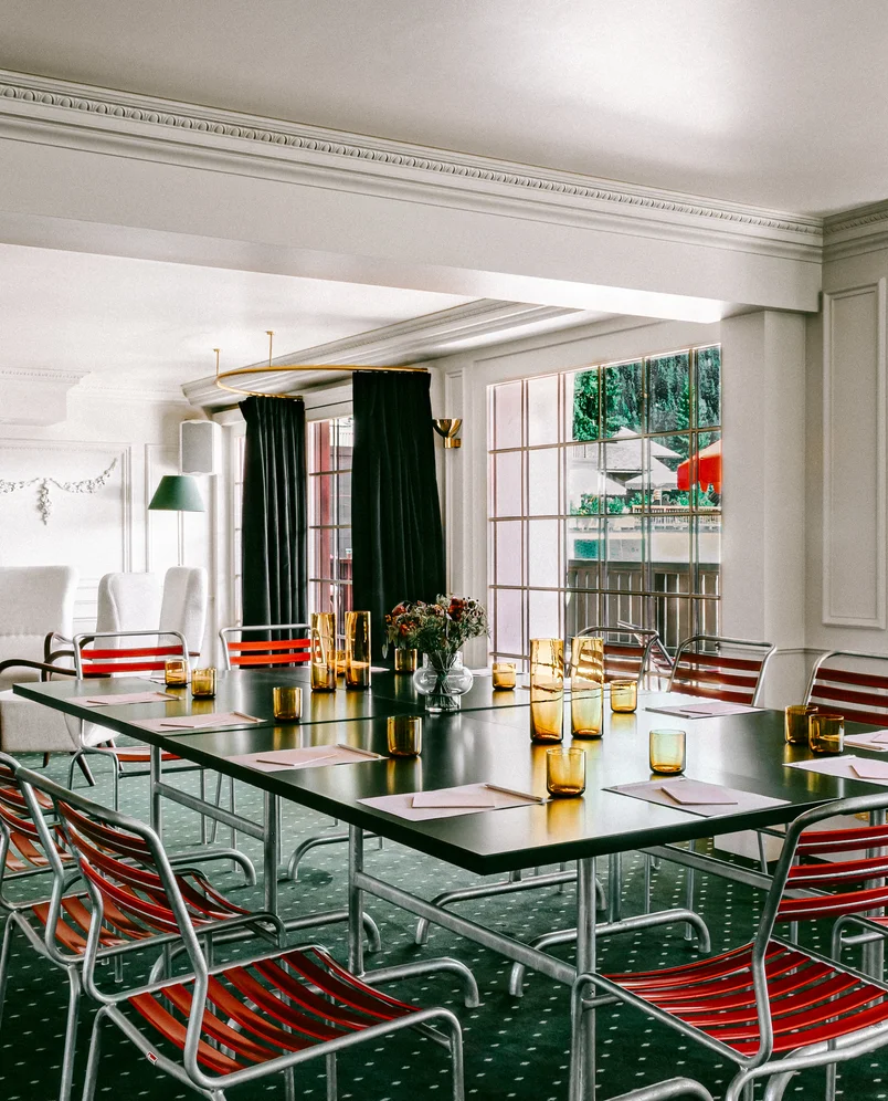 Conference room with black tables arranged in a square, red-striped metal chairs, amber glasses, papers, and flowers, with large windows and green carpet.