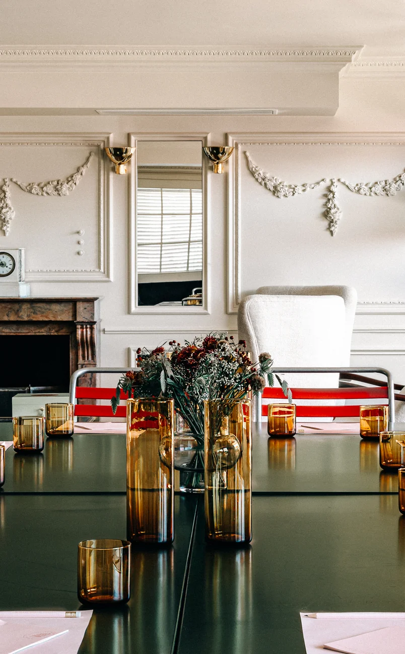 Modern meeting room with a black table set with amber glassware, floral centerpiece, and a mix of red and white chairs against a classic white wall with decorative molding and a mirror.
