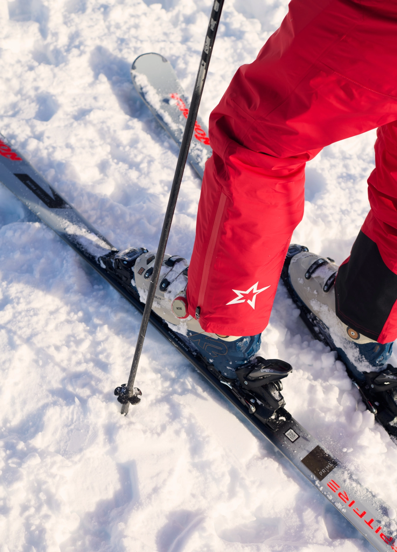 Person wearing red ski pants and ski boots standing on skis in snow with a ski pole planted.