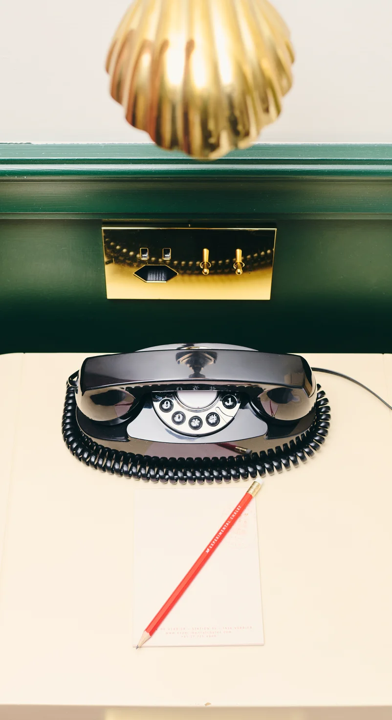 Vintage black rotary telephone on a cream-colored desk with a red pencil and notepad, and a golden lamp above.