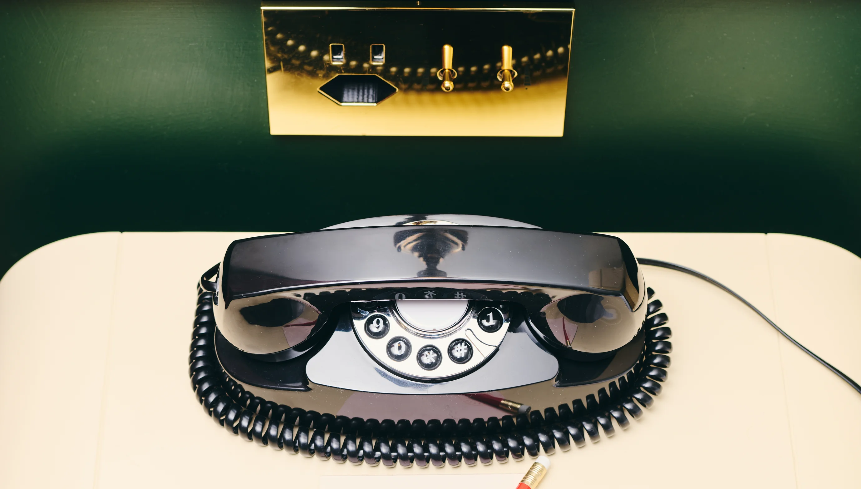 Black rotary dial telephone with coiled cord on a beige surface and gold wall switches above.