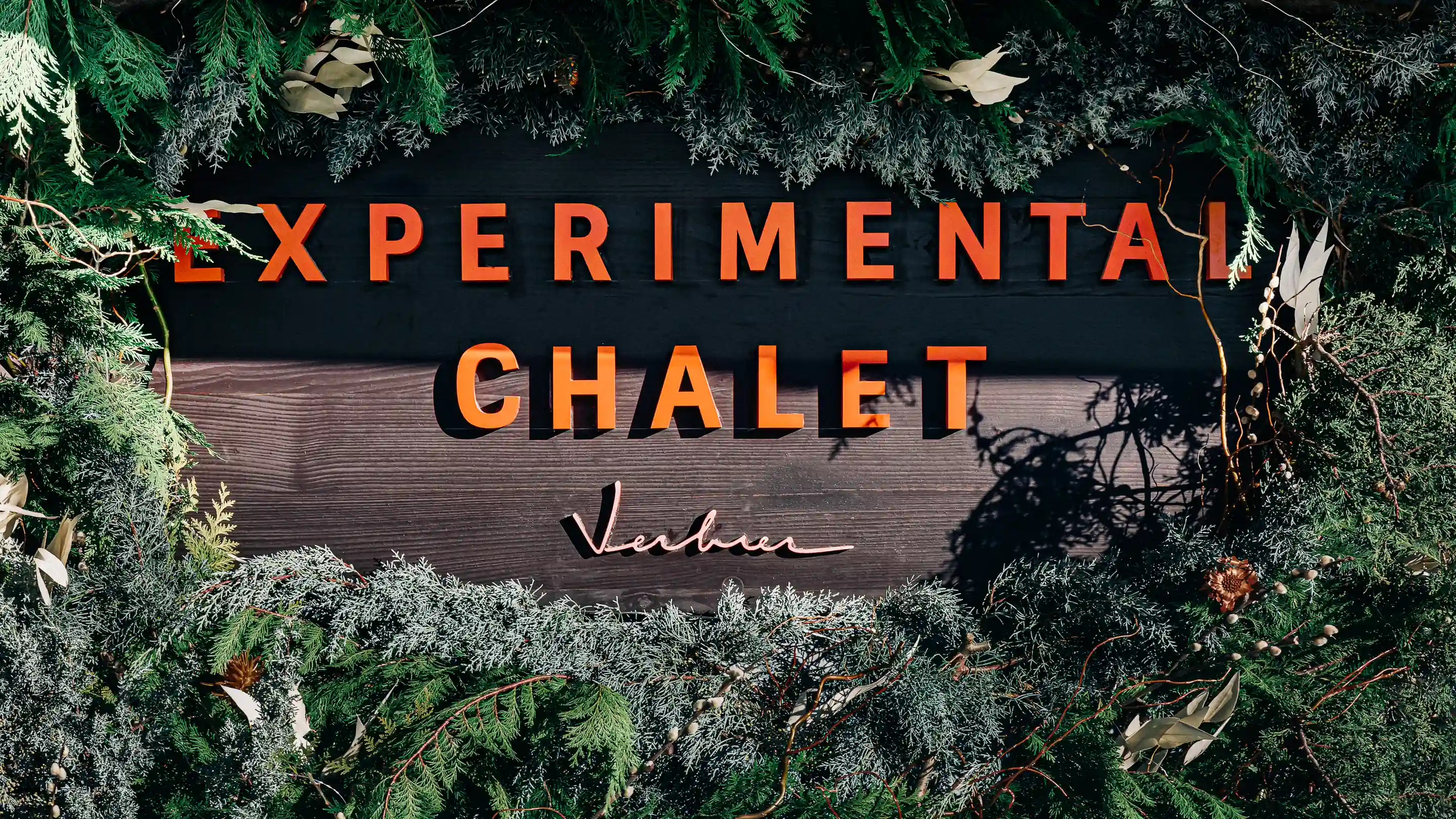 Wooden sign with orange letters reading 'EXPERIMENTAL CHALET Verbier' surrounded by green foliage and pine cones.