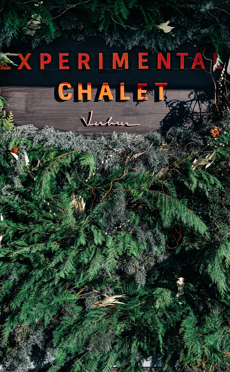 Wooden sign reading 'EXPERIMENTAL CHALET Verbier' surrounded by green pine and fir foliage.