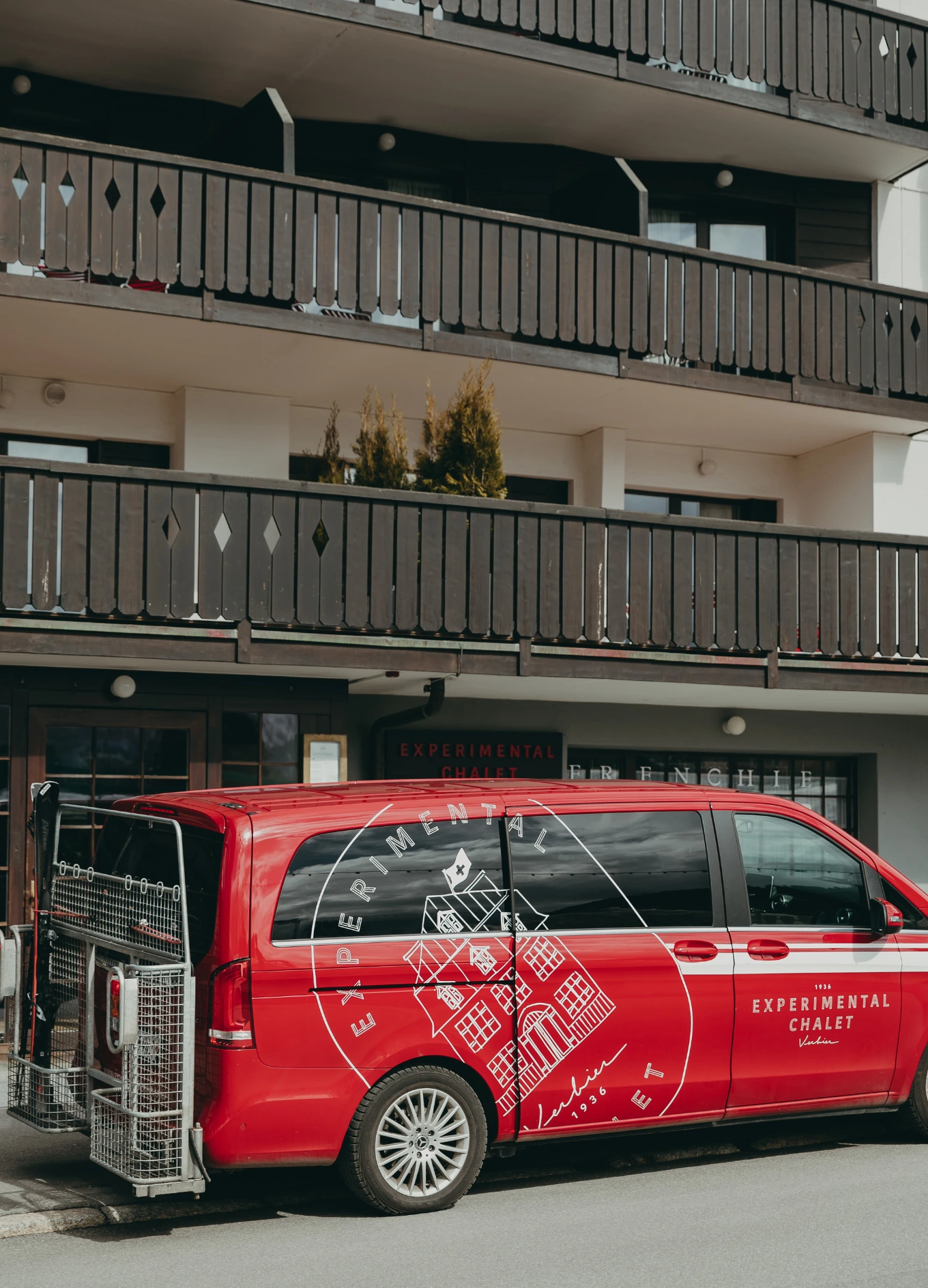 Red van with Experimental Chalet logo parked in front of a building with wooden balconies.