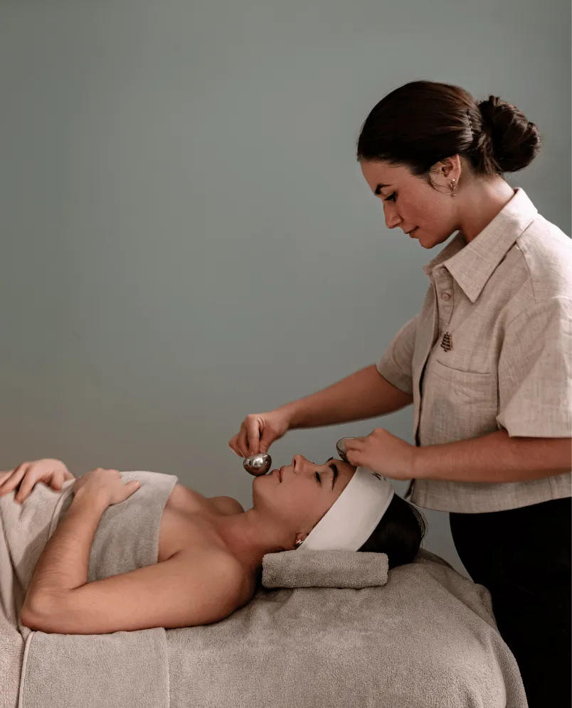 Therapist performing a facial massage on a woman lying on a spa bed with a towel and headband.