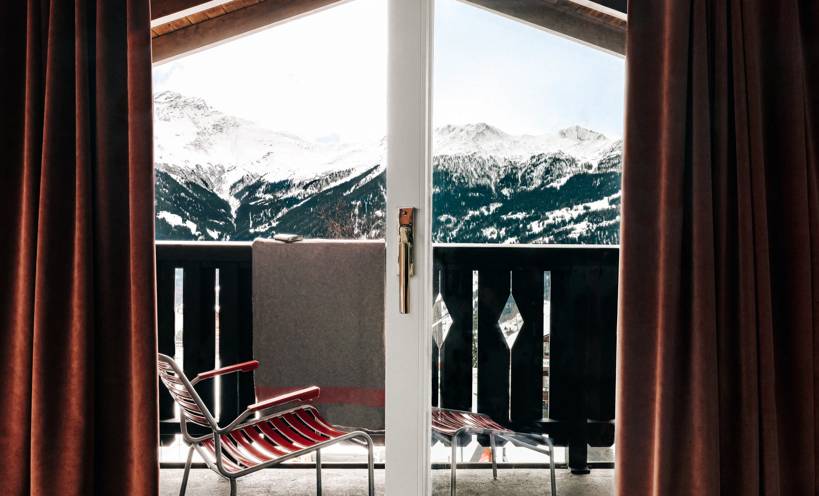 Balcony with metal chair and blanket overlooking snow-covered mountains framed by brown curtains.