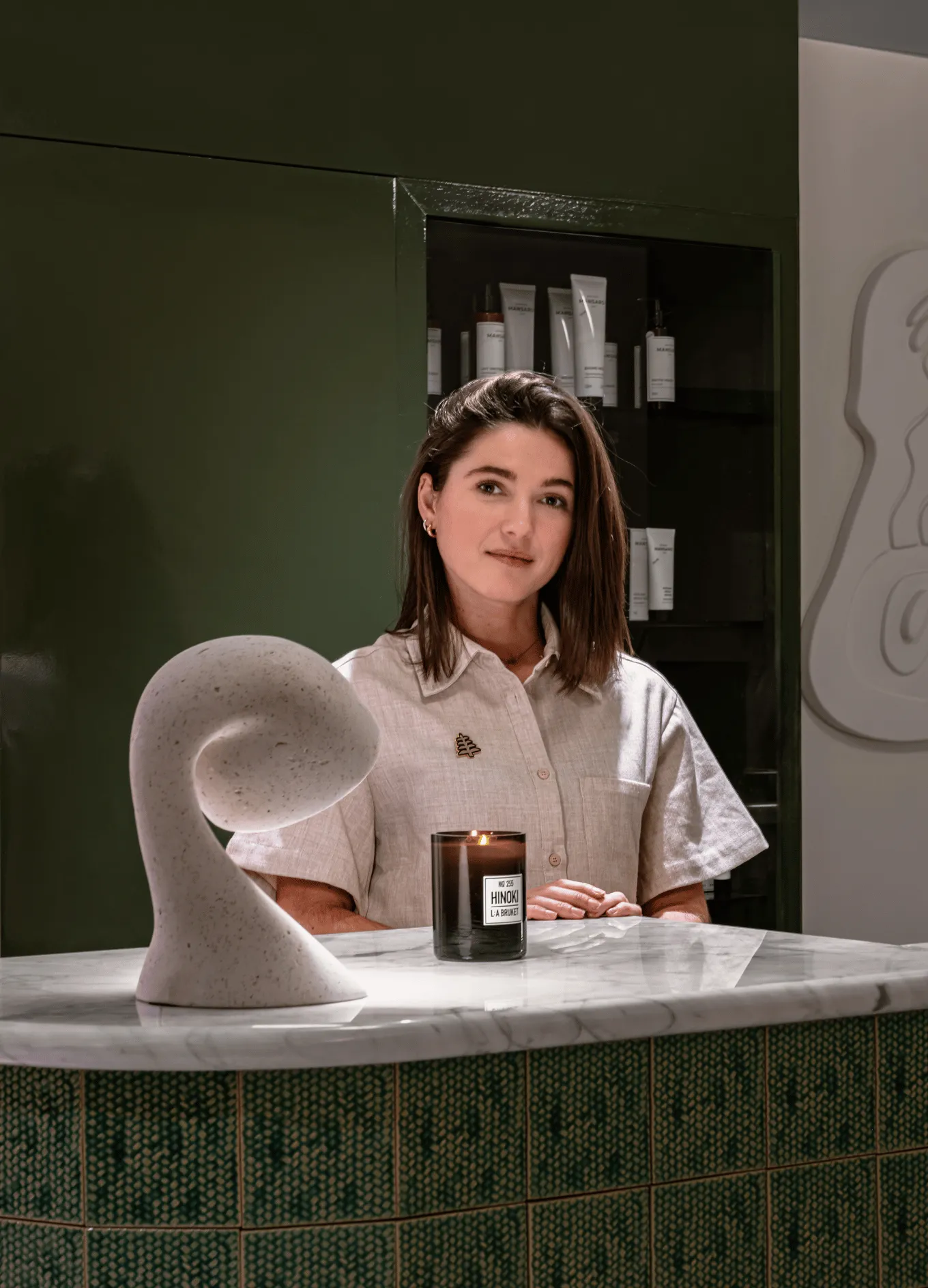 Woman standing behind a marble reception desk with a lit Hinoki scented candle and modern sculpture, with skincare products in the background.