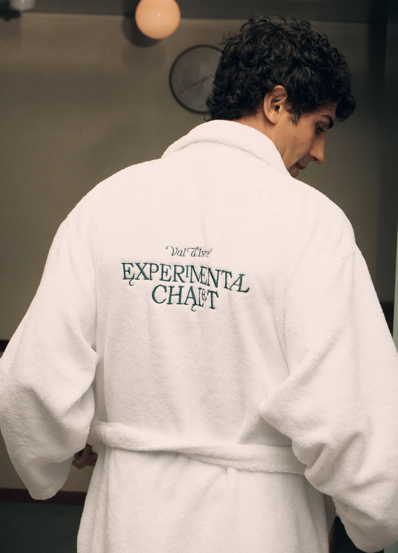 Man wearing a white bathrobe with 'Val d'Isere Experimental Chalet' embroidered on the back in a dimly lit room.