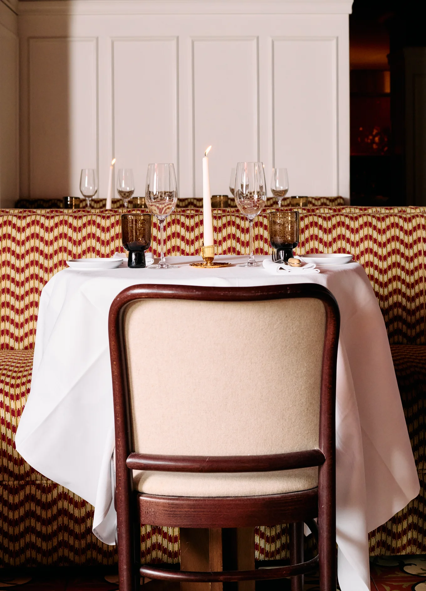 Table set for two with white tablecloth, wine glasses, brown water glasses, plates, napkins, and a lit candle in an elegant restaurant.