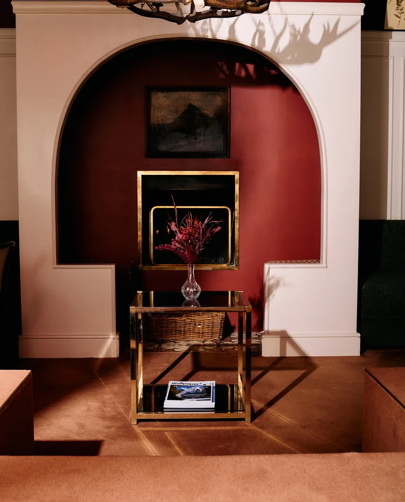 Modern interior with an arched alcove featuring a dark red wall, a glass vase with red flowers on a reflective gold-framed table, and a magazine on the lower shelf.