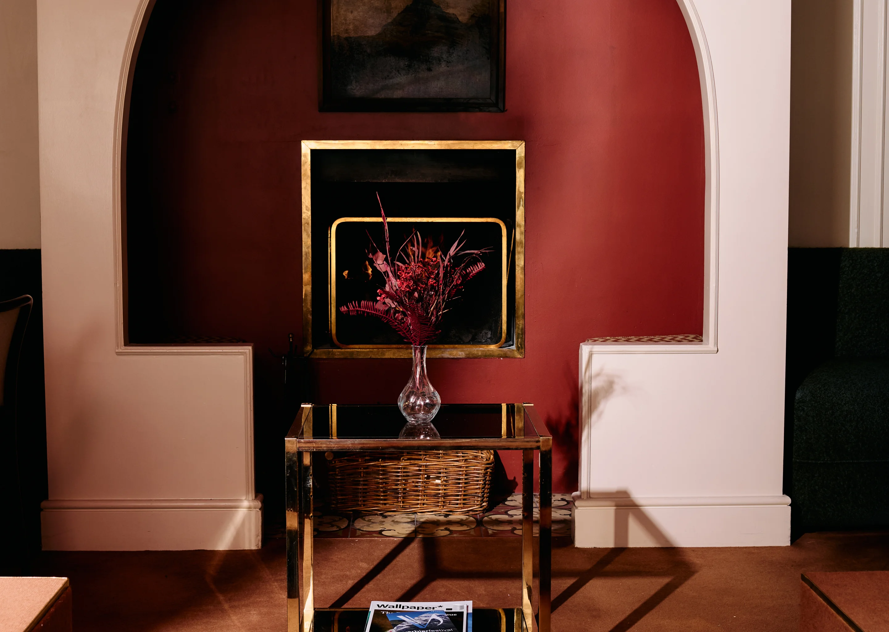 Modern living room with a brass-framed fireplace set in a red wall alcove, a glass coffee table with a vase of dried red flowers, and a wicker basket underneath.