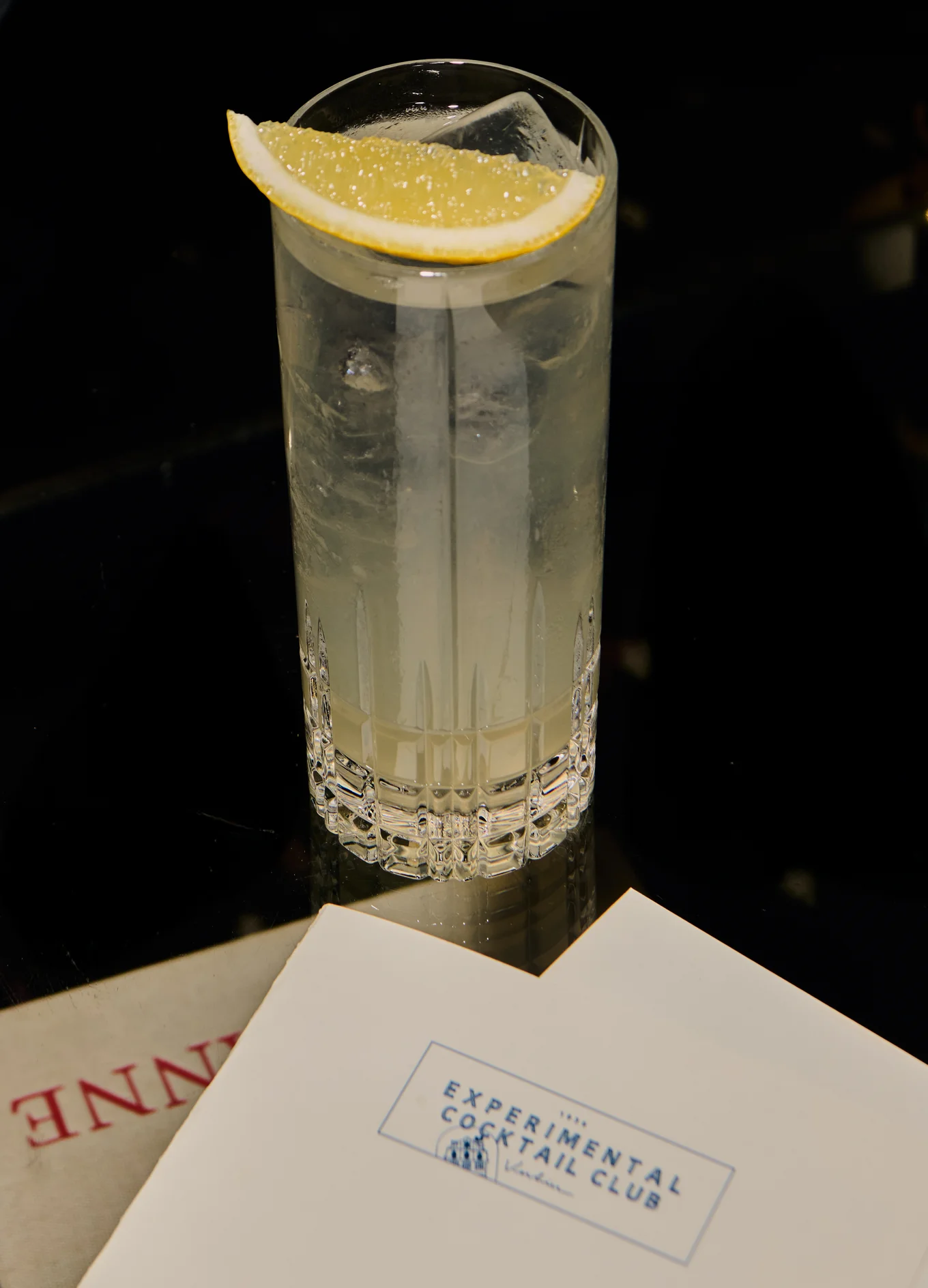 Tall crystal glass with a lemon wedge and ice cubes containing a clear cocktail on a black reflective surface next to a card labeled Experimental Cocktail Club.