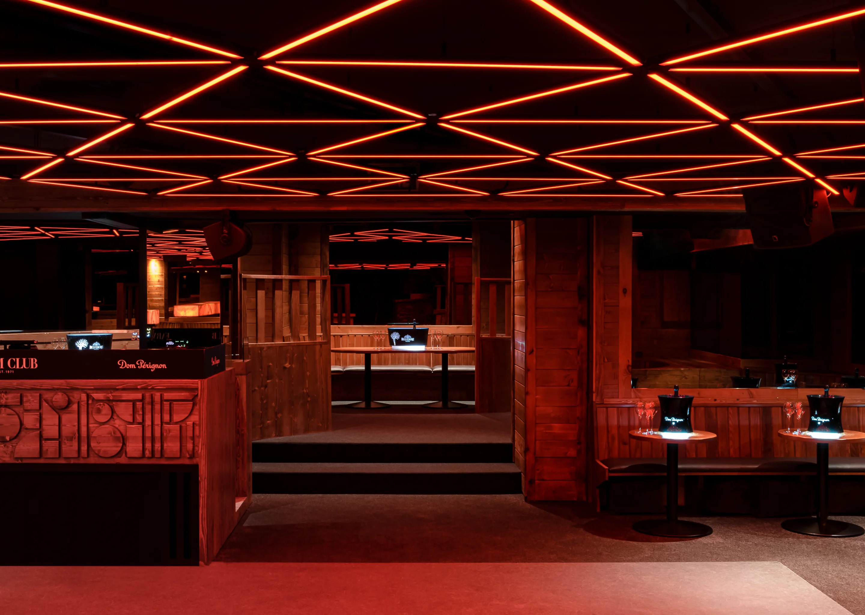 Interior of a modern lounge with geometric red ceiling lights, wooden paneling, tables with Dom Pérignon buckets, and seating areas.