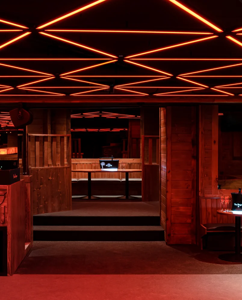 Dimly lit interior of a lounge with wooden paneling, steps, and red neon geometric ceiling lights.