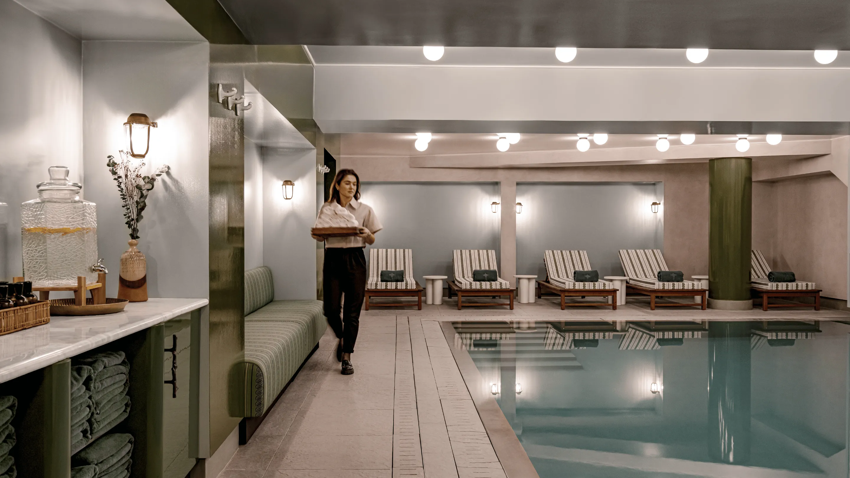 Indoor wellness pool area with striped lounge chairs, green towels, soft lighting, and a woman carrying a tray of rolled towels.