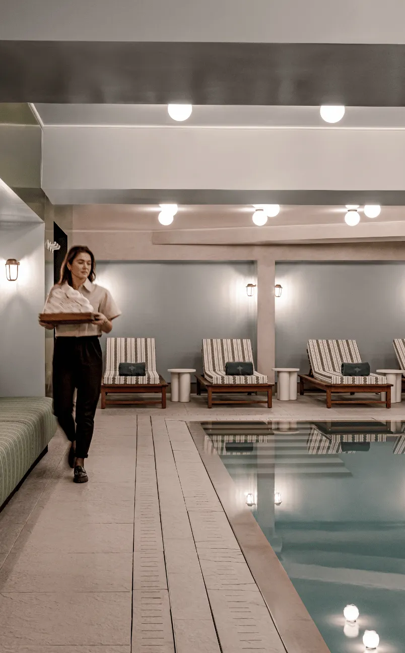 Woman in uniform carrying a tray of towels walking beside an indoor pool with lounge chairs and soft lighting.