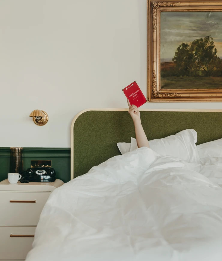 Person lying in bed with white bedding, holding up a red Après-Ski Map booklet next to a green headboard and vintage painting on the wall.