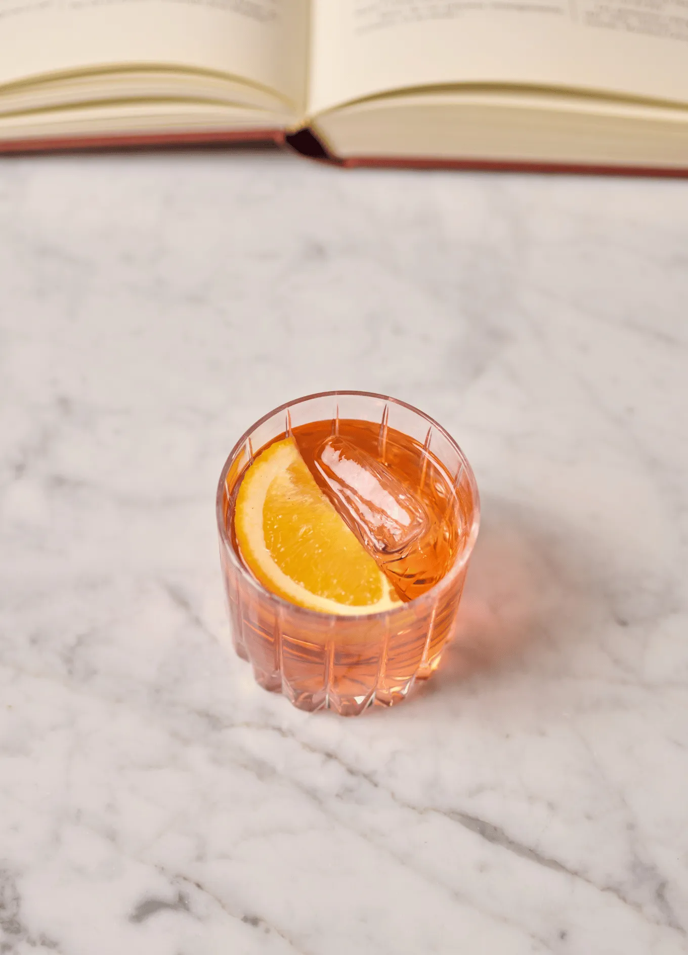 Glass of orange-colored drink with ice and an orange slice on a marble surface, with an open book in the background.