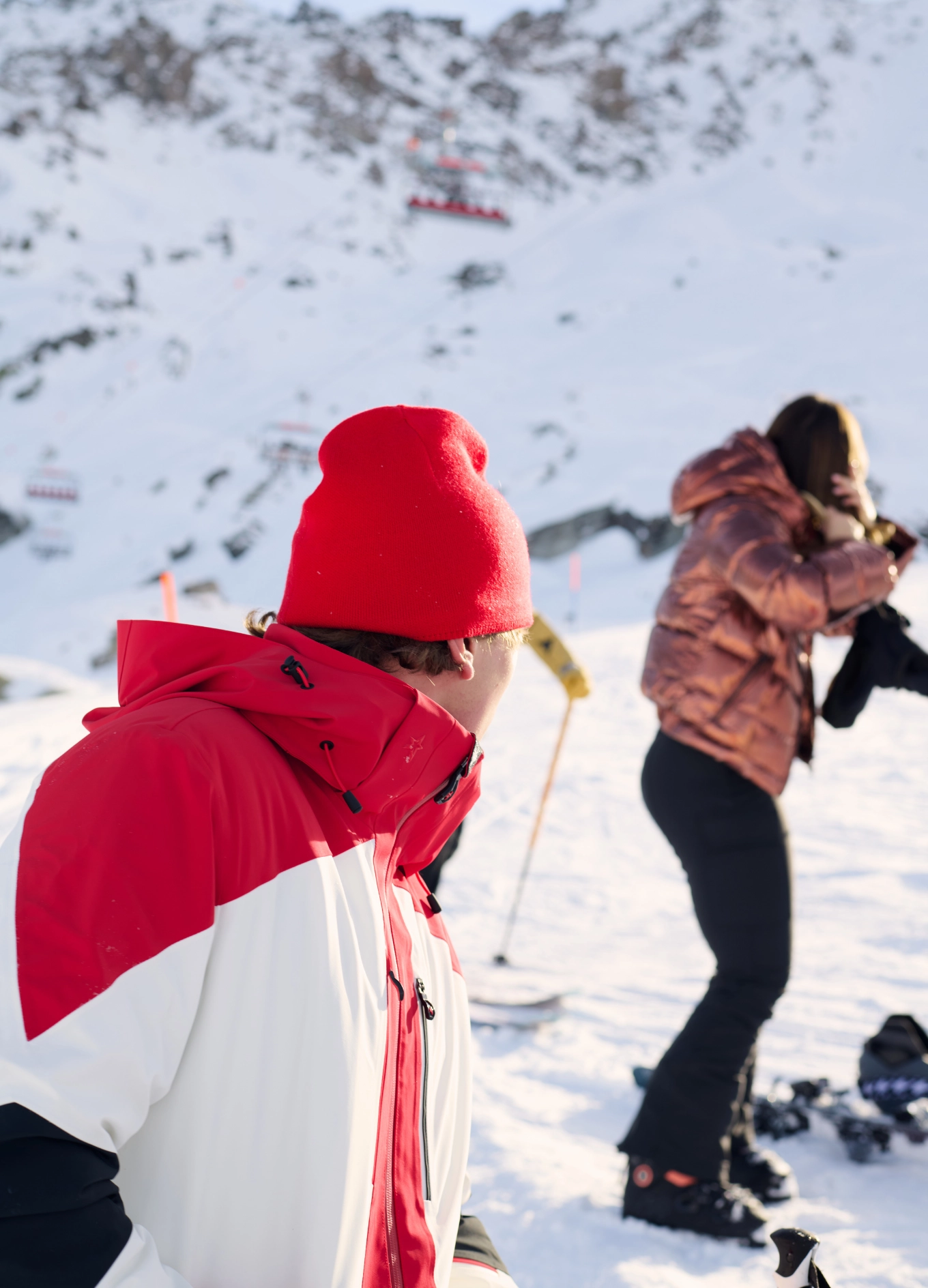Person in a red and white ski jacket and red beanie looking at another person adjusting their hair on a snowy mountain slope.