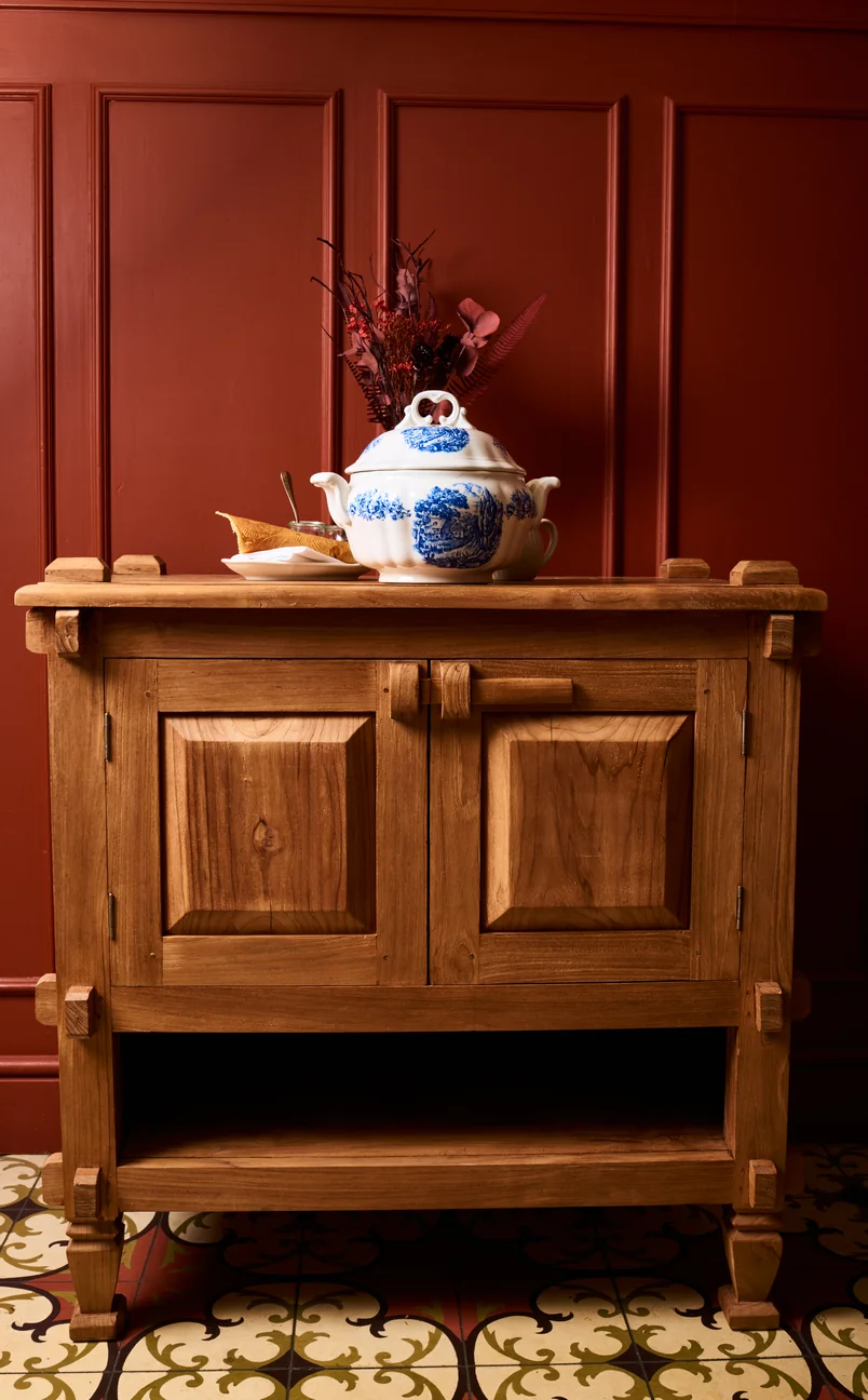 Wooden cabinet with a white and blue porcelain soup tureen, a plate, and dried flowers on top against a dark red paneled wall.