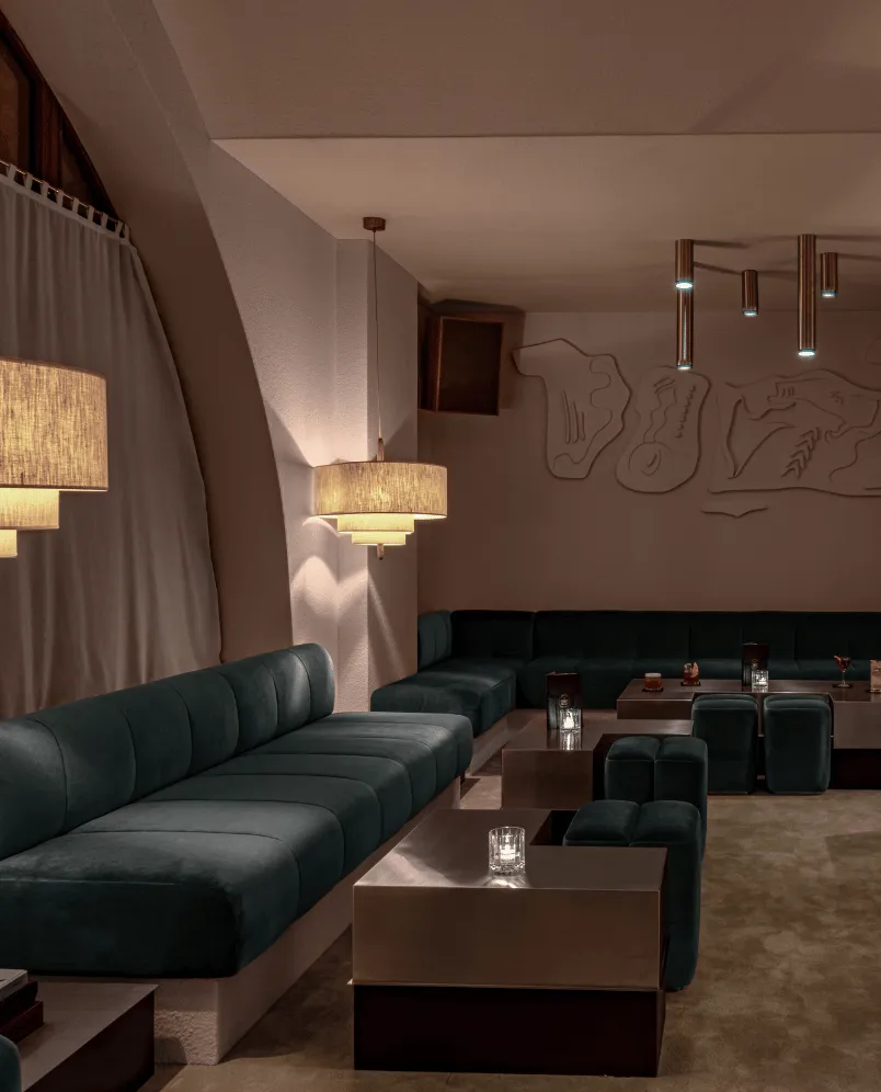 Modern lounge with dark green plush sofas, low square tables, pendant lamps, and abstract wall art.
