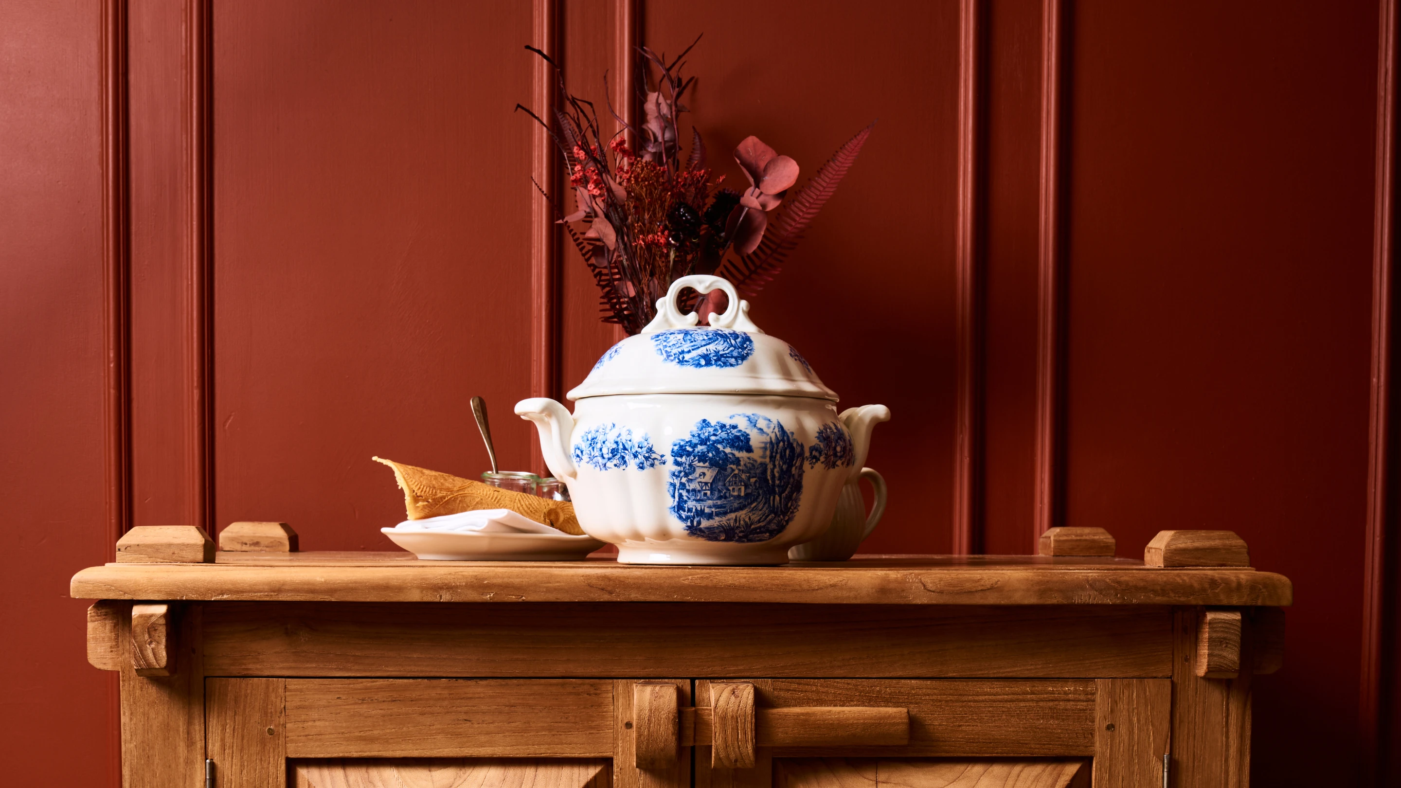 Blue and white porcelain tureen with floral and cottage design on wooden cabinet against a red paneled wall with dried flowers in the background.